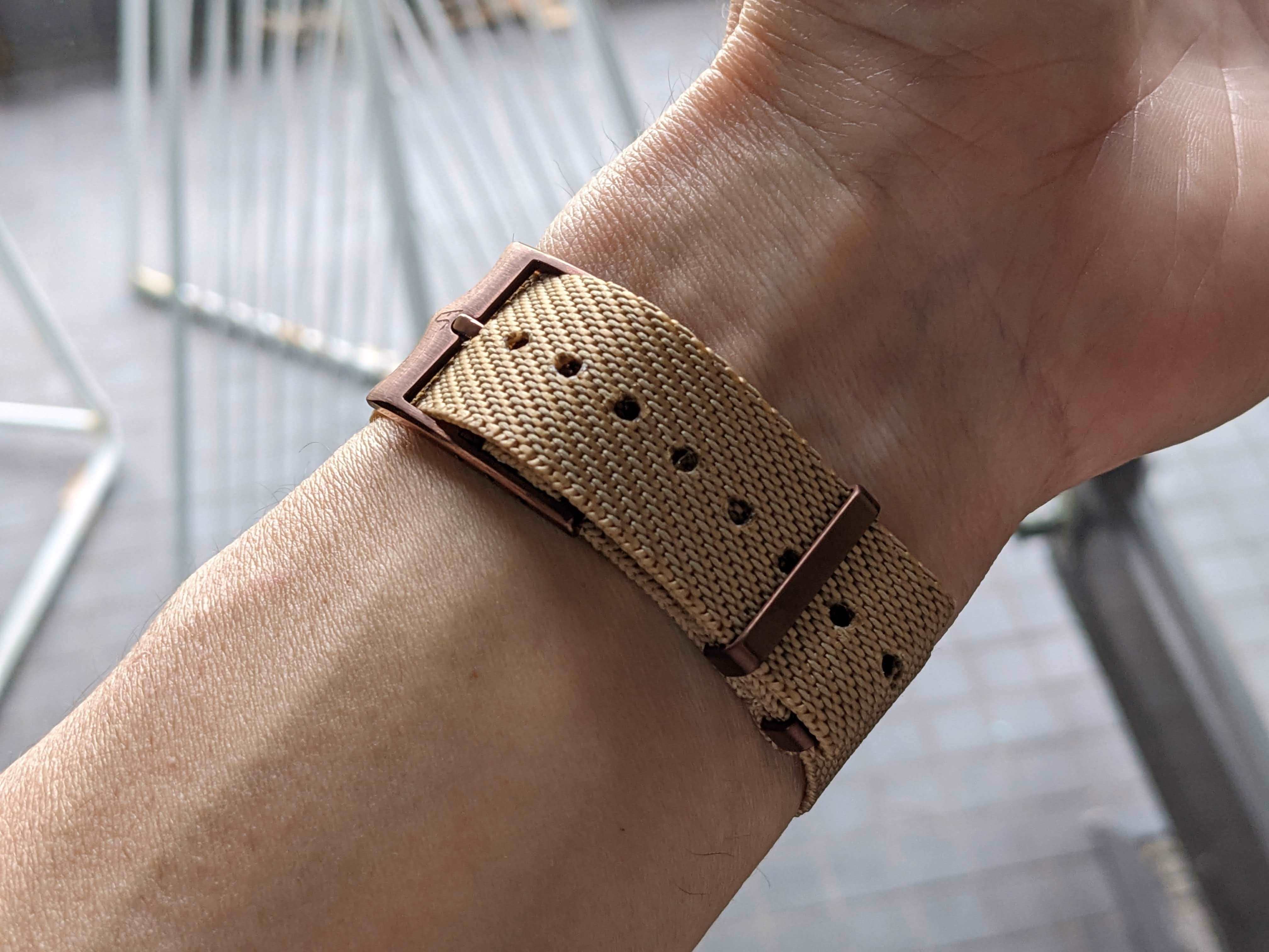 Tan canvas watch strap with bronze buckle worn on wrist, adjustable military-style band with perforated holes