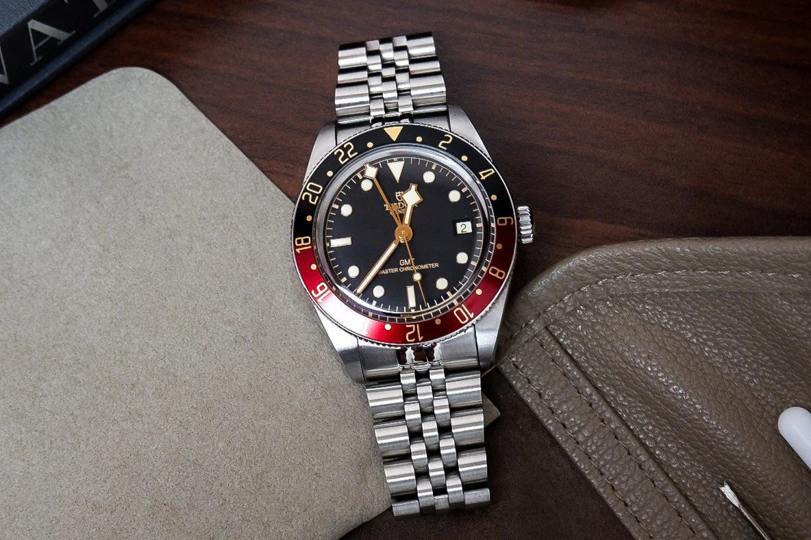 Luxury steel sports watch with red and black GMT bezel, black dial, applied indices, and matching jubilee bracelet on wooden desk