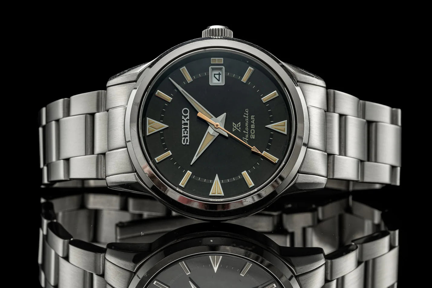 Seiko Alpinist stainless steel watch with black dial, gold hour markers, date window, and matching metal bracelet on reflective surface