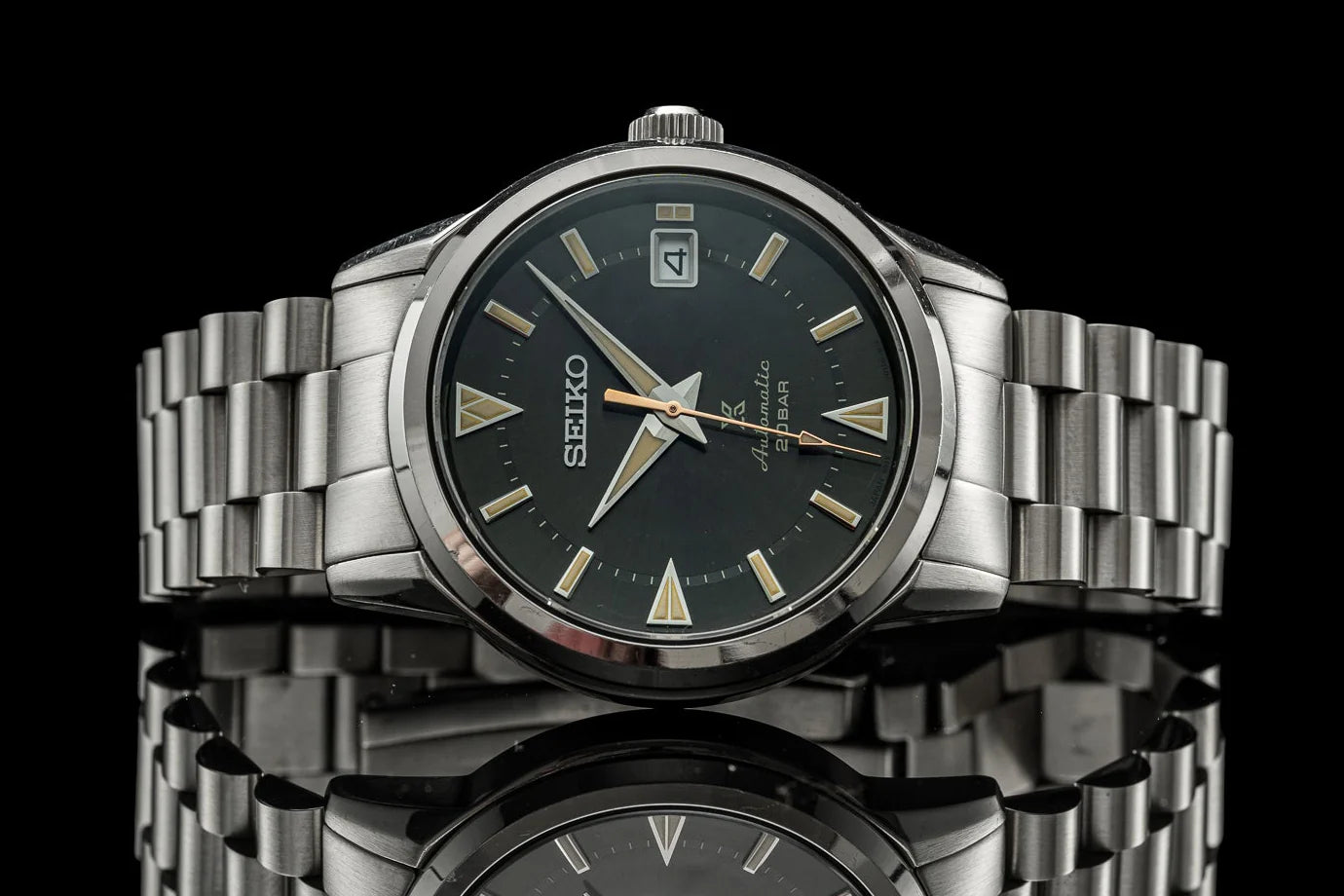 Seiko Alpinist stainless steel watch with black dial, gold indices, and matching steel bracelet on reflective surface