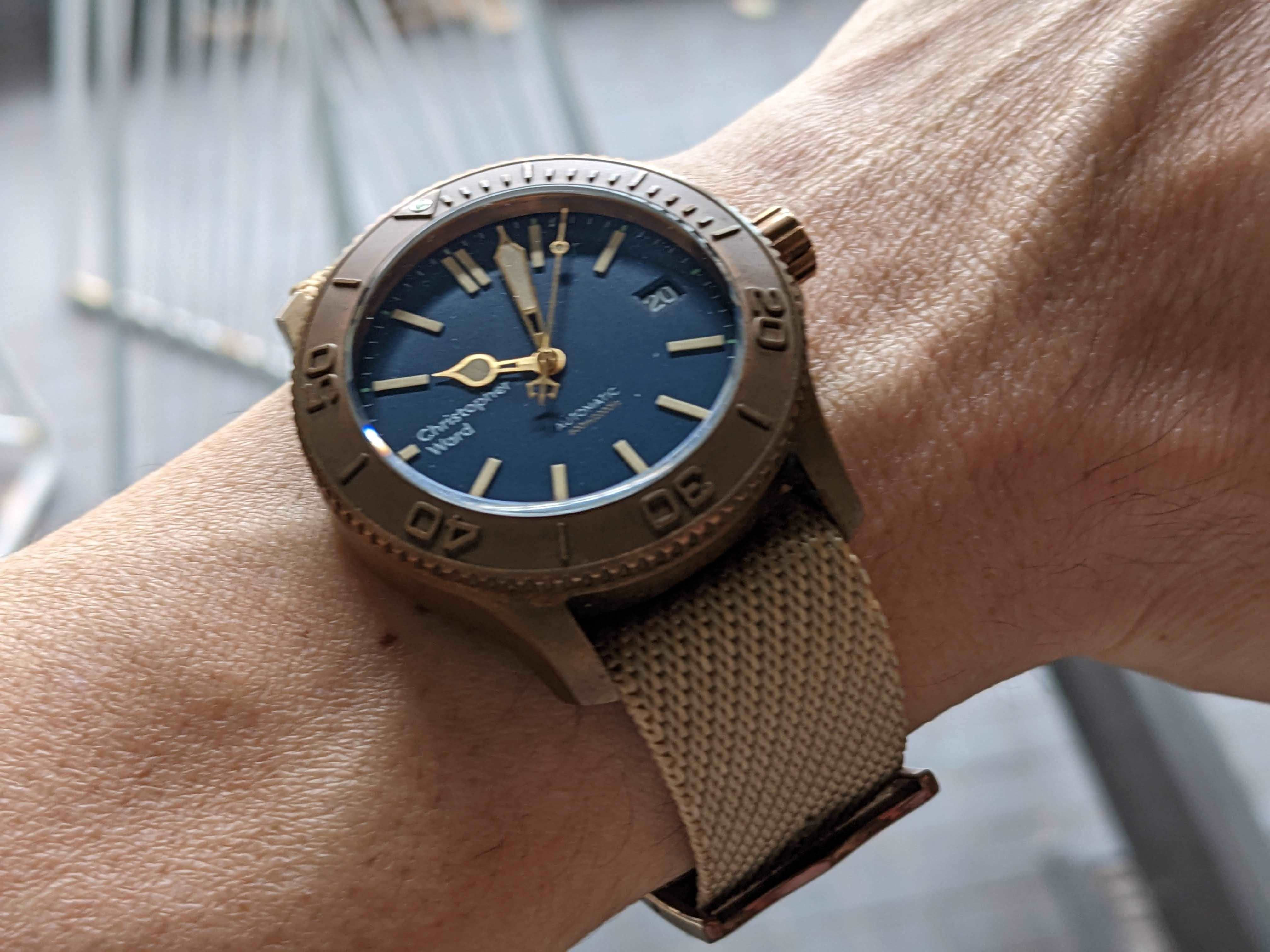 Bronze military watch with blue dial and tan fabric strap worn on wrist, showing adjustable design and tactical styling