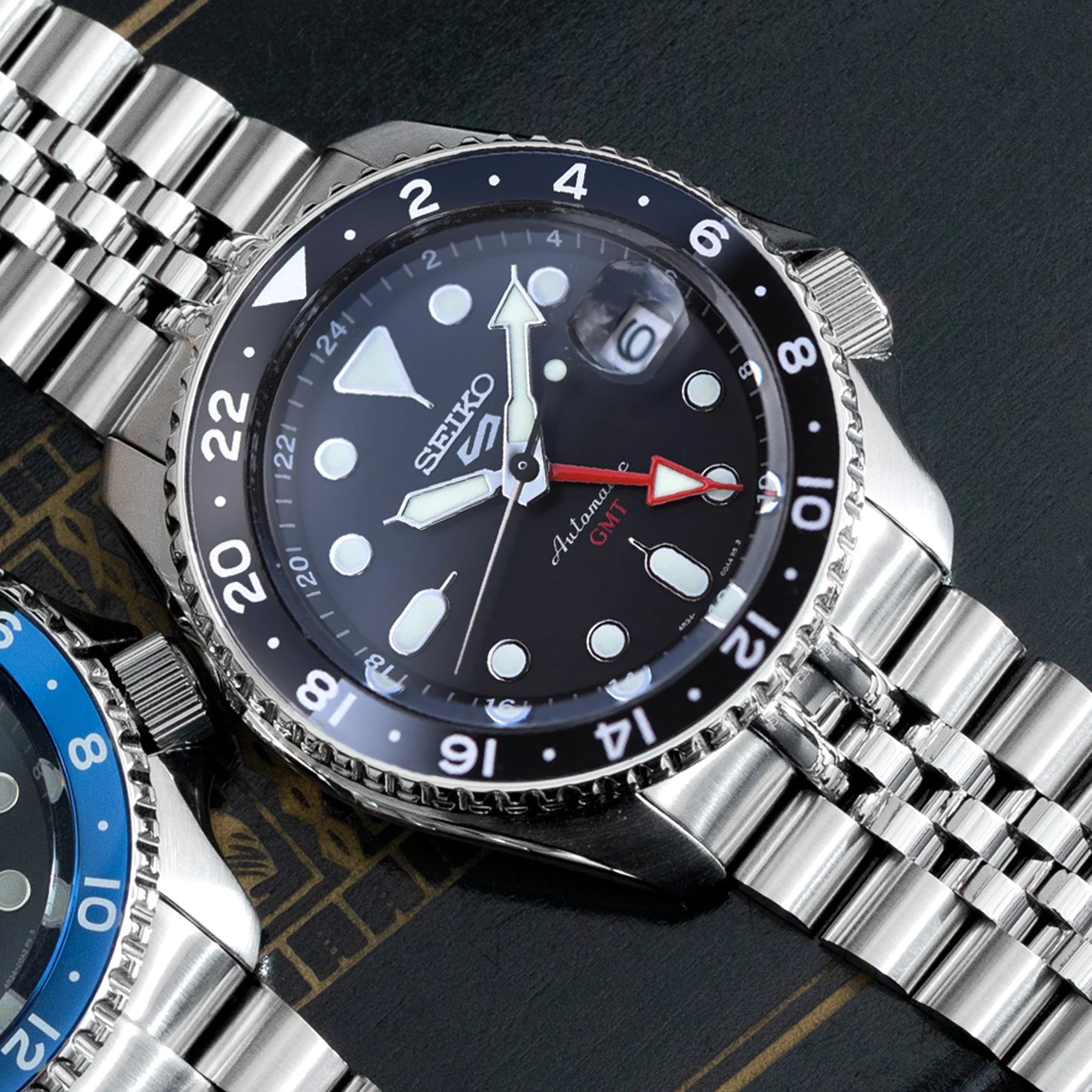 Seiko 5 GMT automatic watch with black dial, steel Super-J Louis bracelet, GMT hand, and rotating bezel on dark background