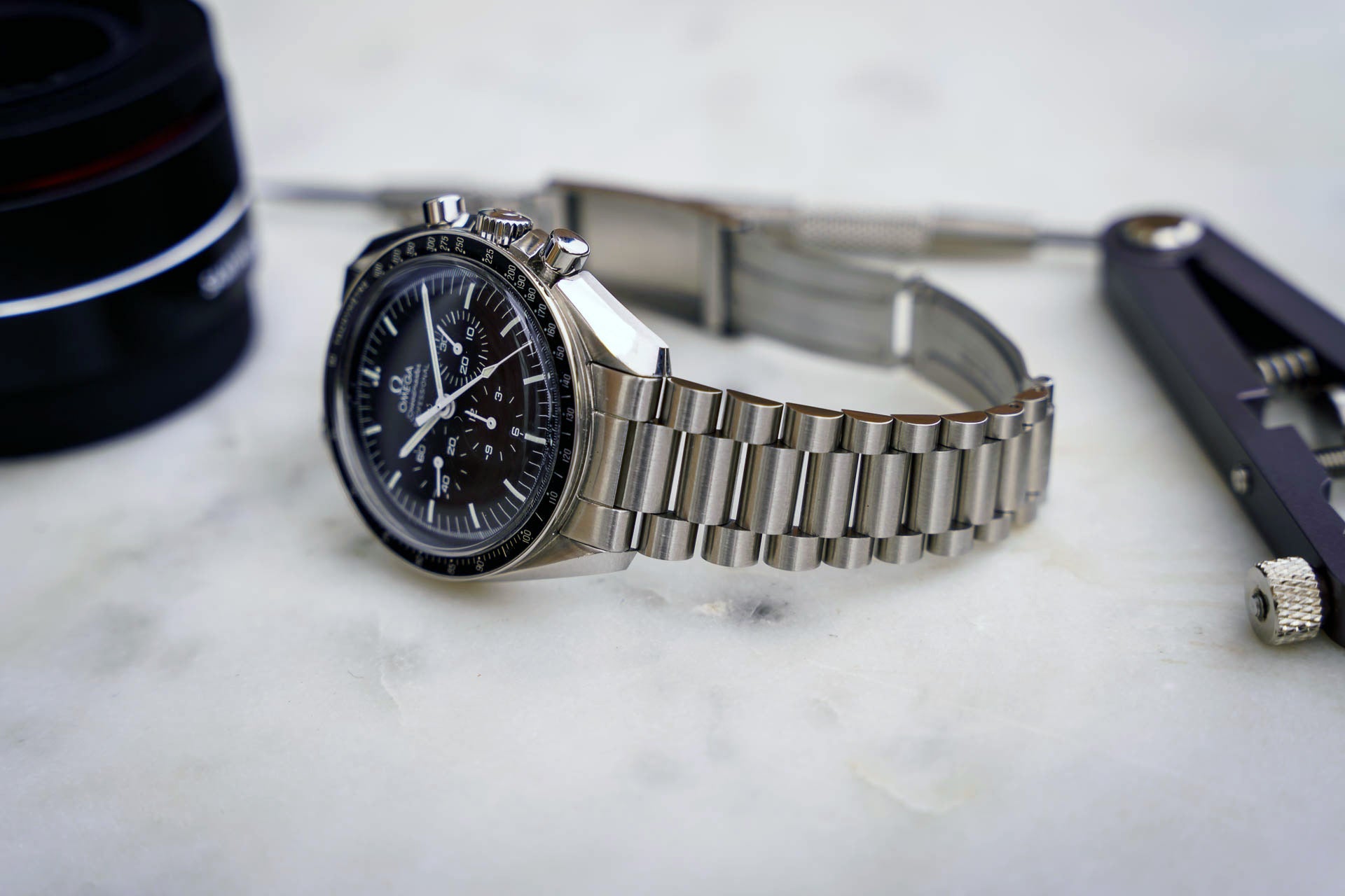 Stainless steel chronograph watch with black dial and metal bracelet on marble surface alongside accessories