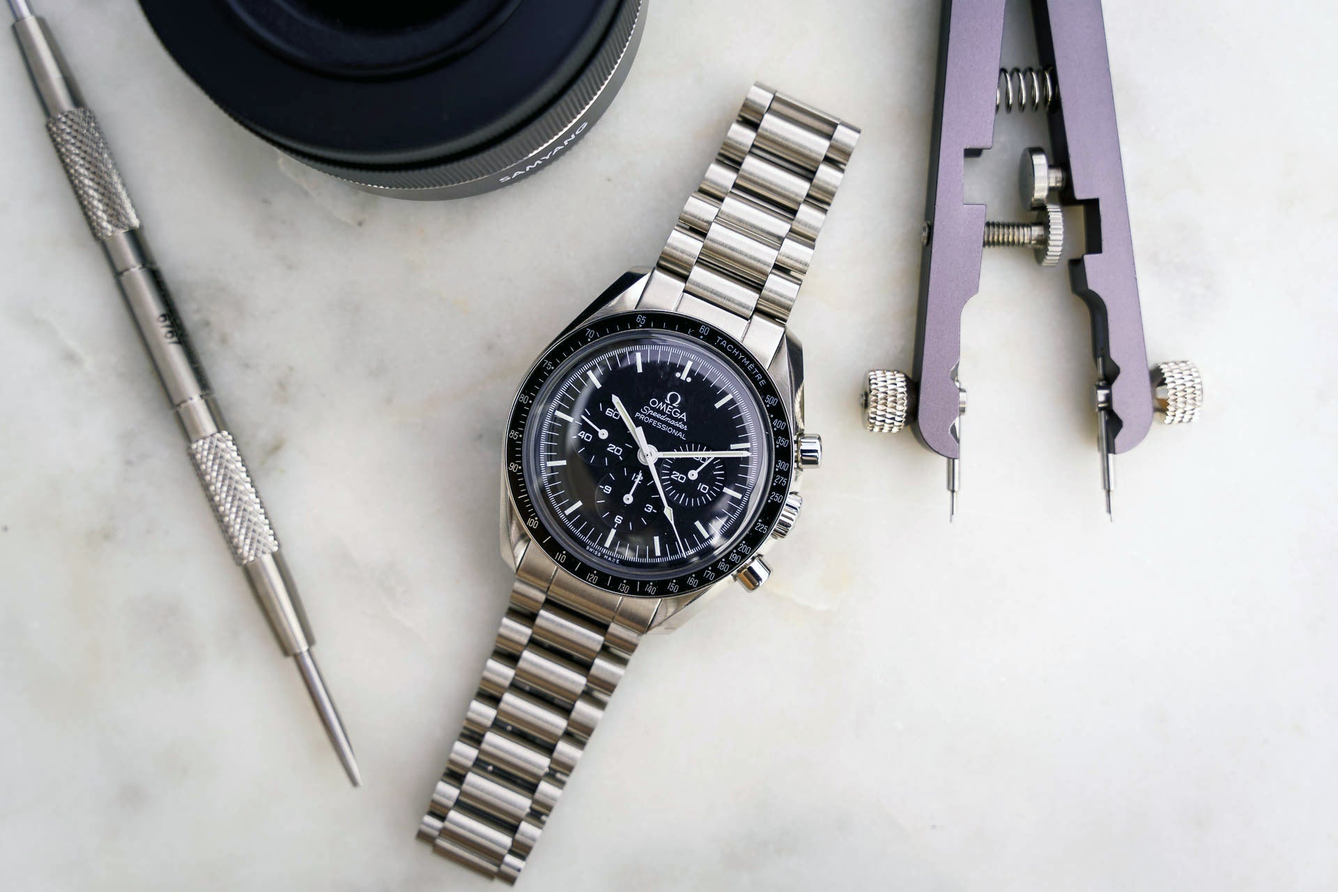 Omega Speedmaster chronograph watch with stainless steel bracelet displayed with watchmaker tools and case on marble surface