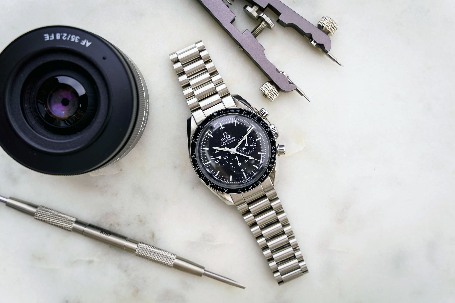 Omega Speedmaster professional chronograph watch with stainless steel bracelet and black dial, displayed with camera lens and watchmaking tools