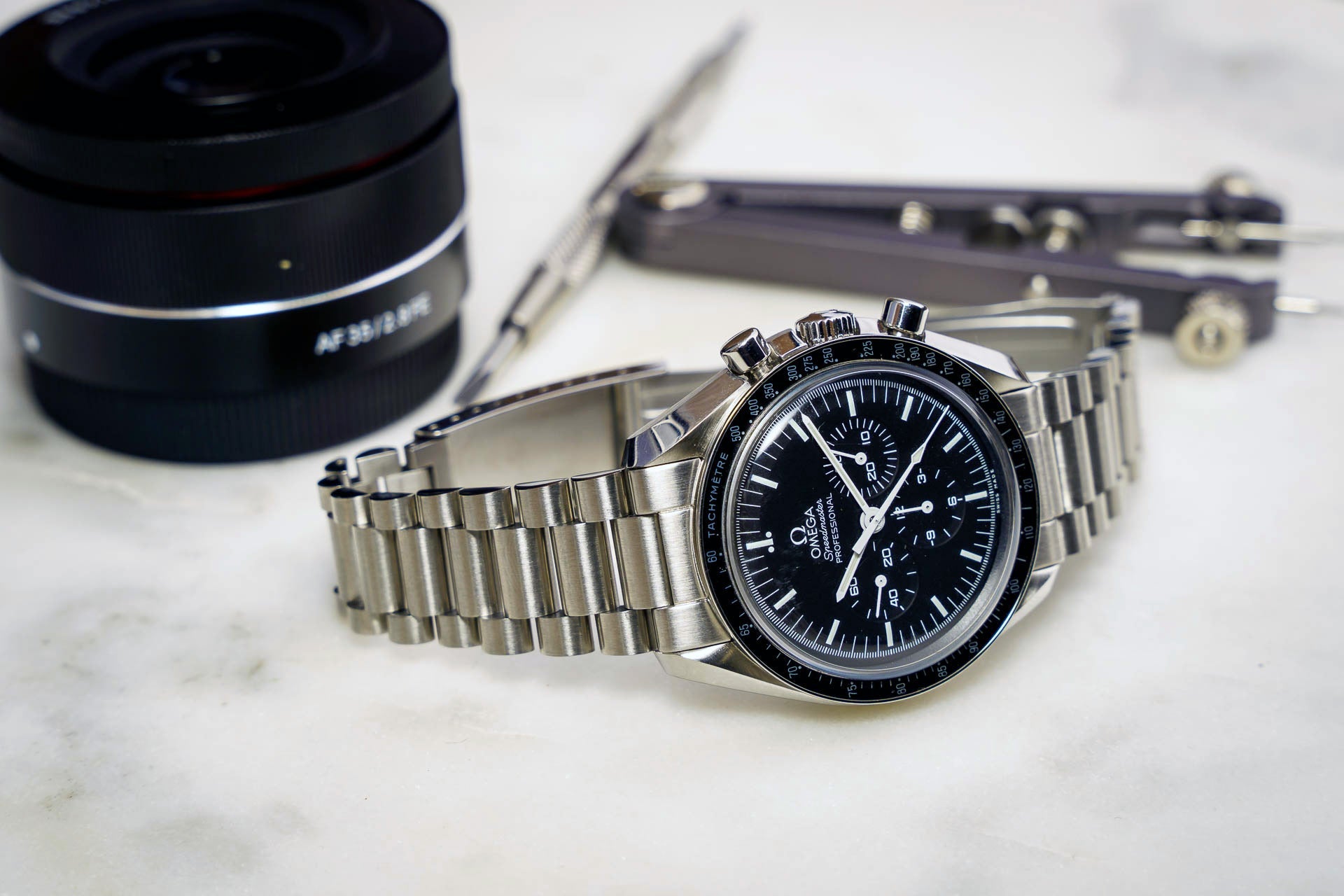 Omega Speedmaster chronograph watch with stainless steel bracelet and black dial displayed with camera lens and accessories