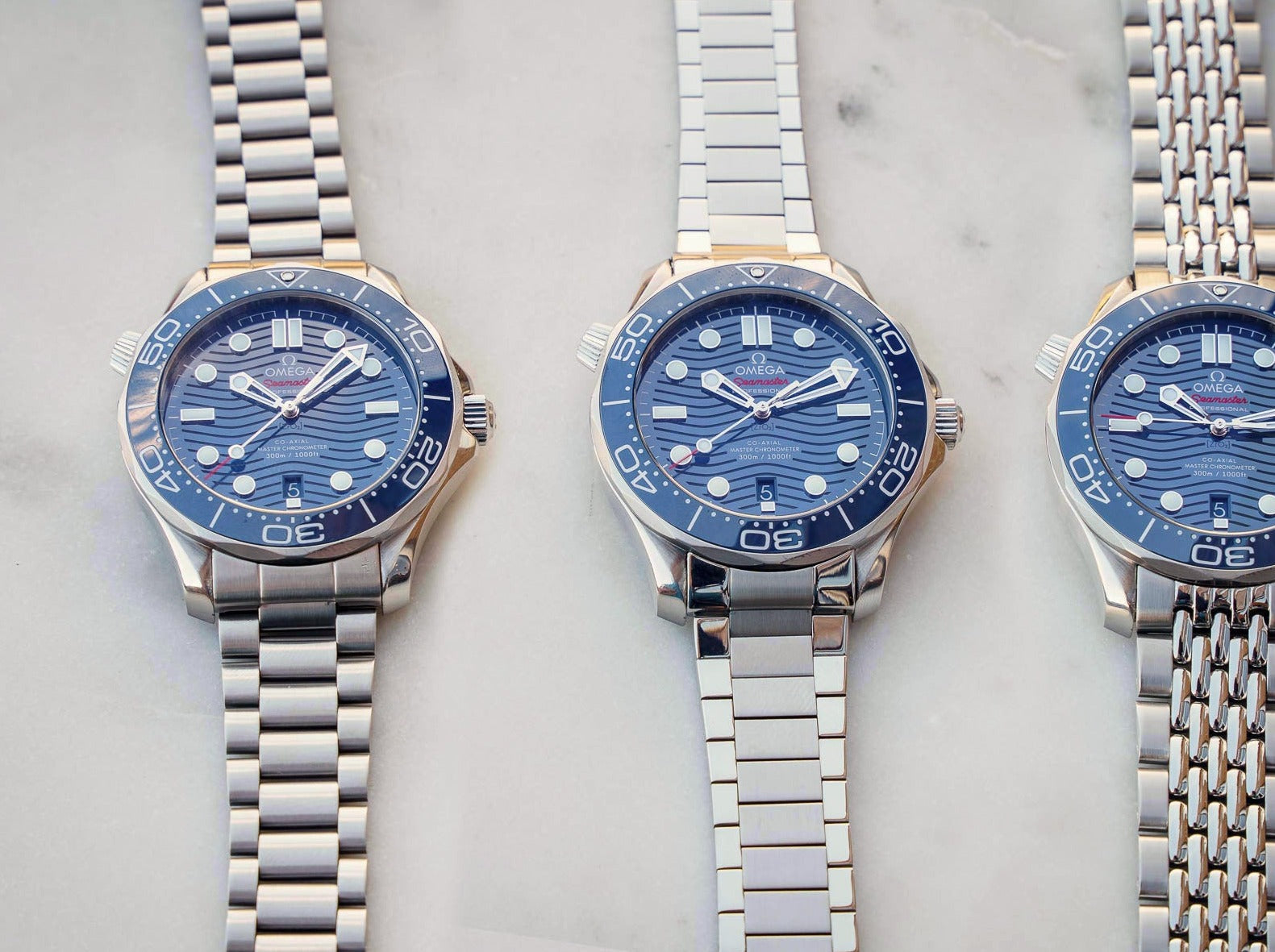 Three Omega Seamaster 300m dive watches with blue wave dials and stainless steel bracelets in different styles on marble background