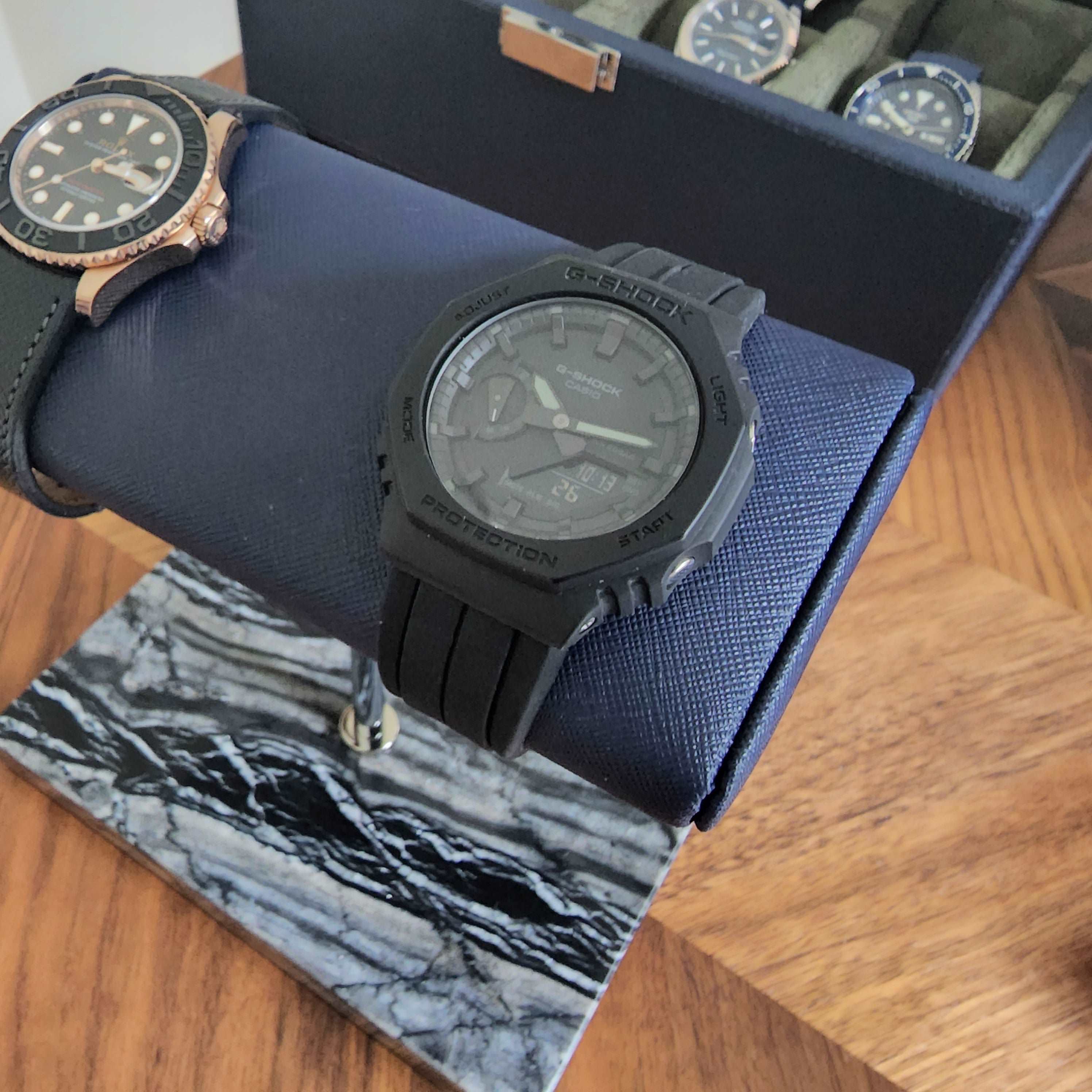 Black G-Shock digital analog watch with silicone band displayed in blue storage box with other timepieces