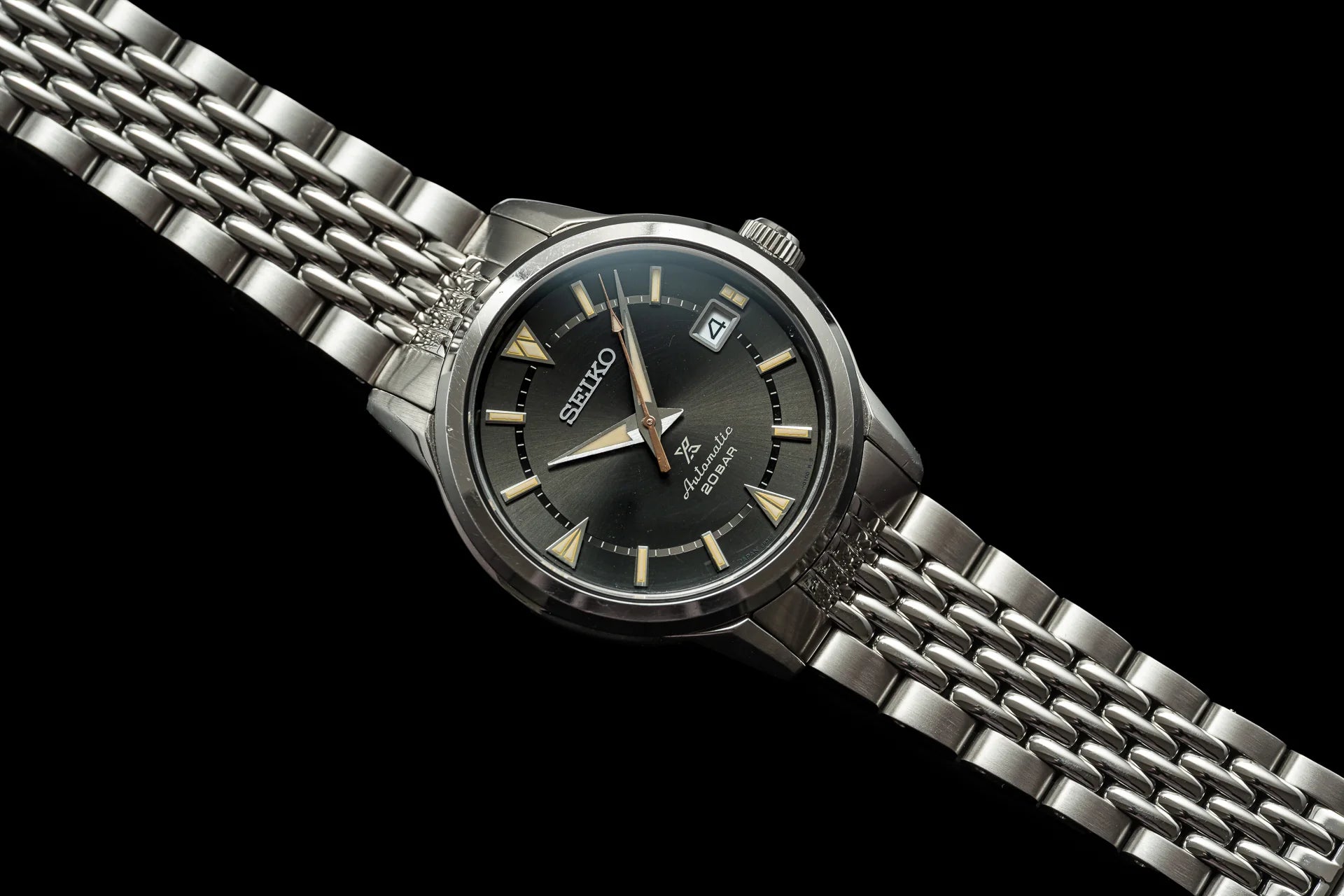 Seiko Alpinist automatic watch with black dial, gold hour markers, date window, and stainless steel beads of rice bracelet on black background