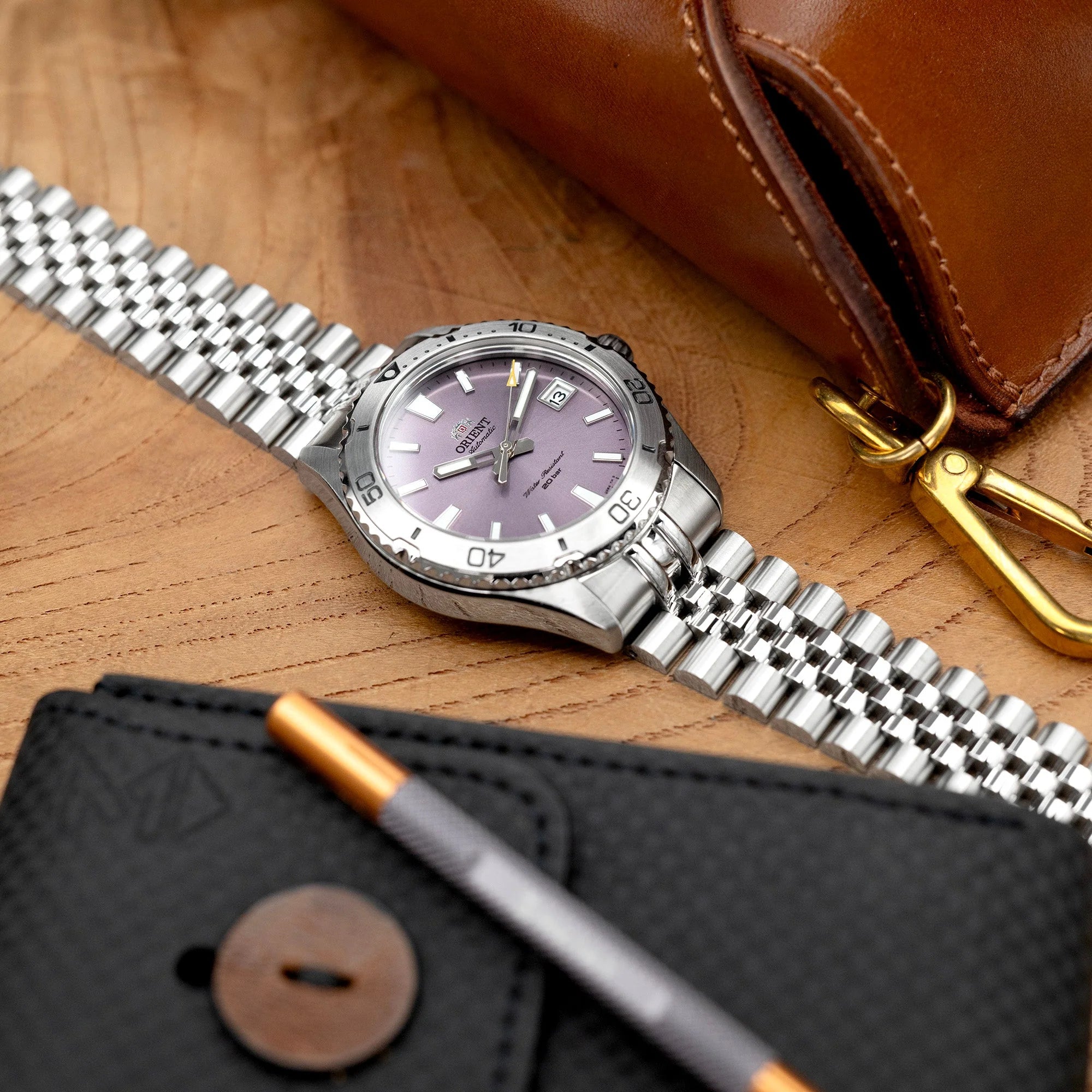Stainless steel Super-JUB II watch band on Orient Mako 40 with purple dial, displayed on wooden surface with brown leather bag