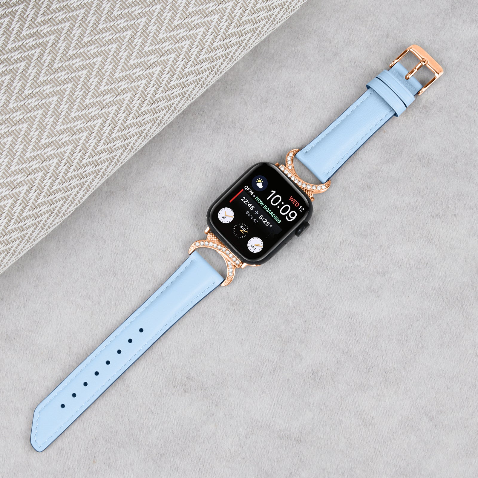 Smartwatch with black square face displaying time 10:09, paired with light blue leather band and rose gold crystal-studded connector lugs