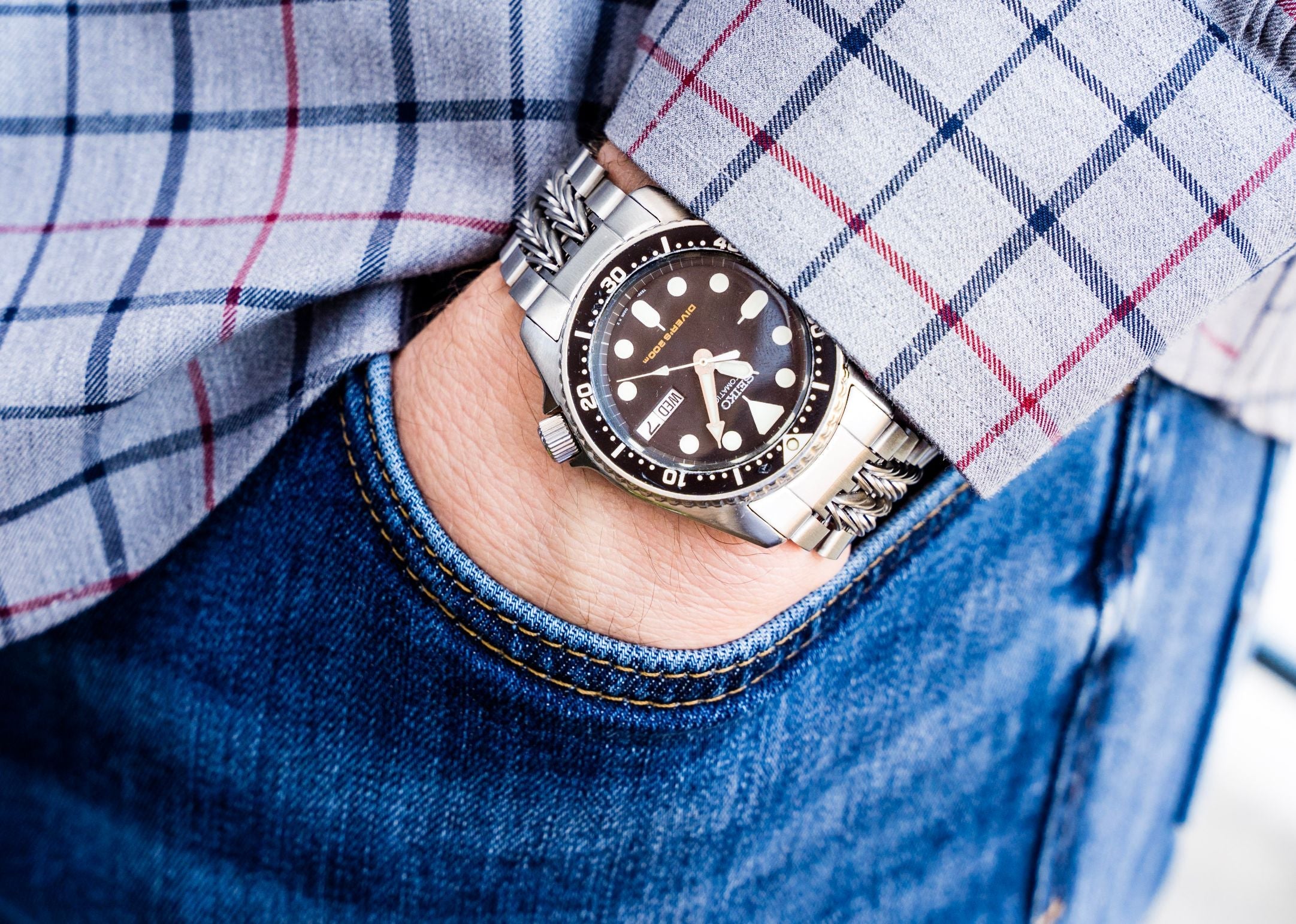 Men's stainless steel dive watch with black dial and dot markers worn on wrist with plaid shirt and jeans
