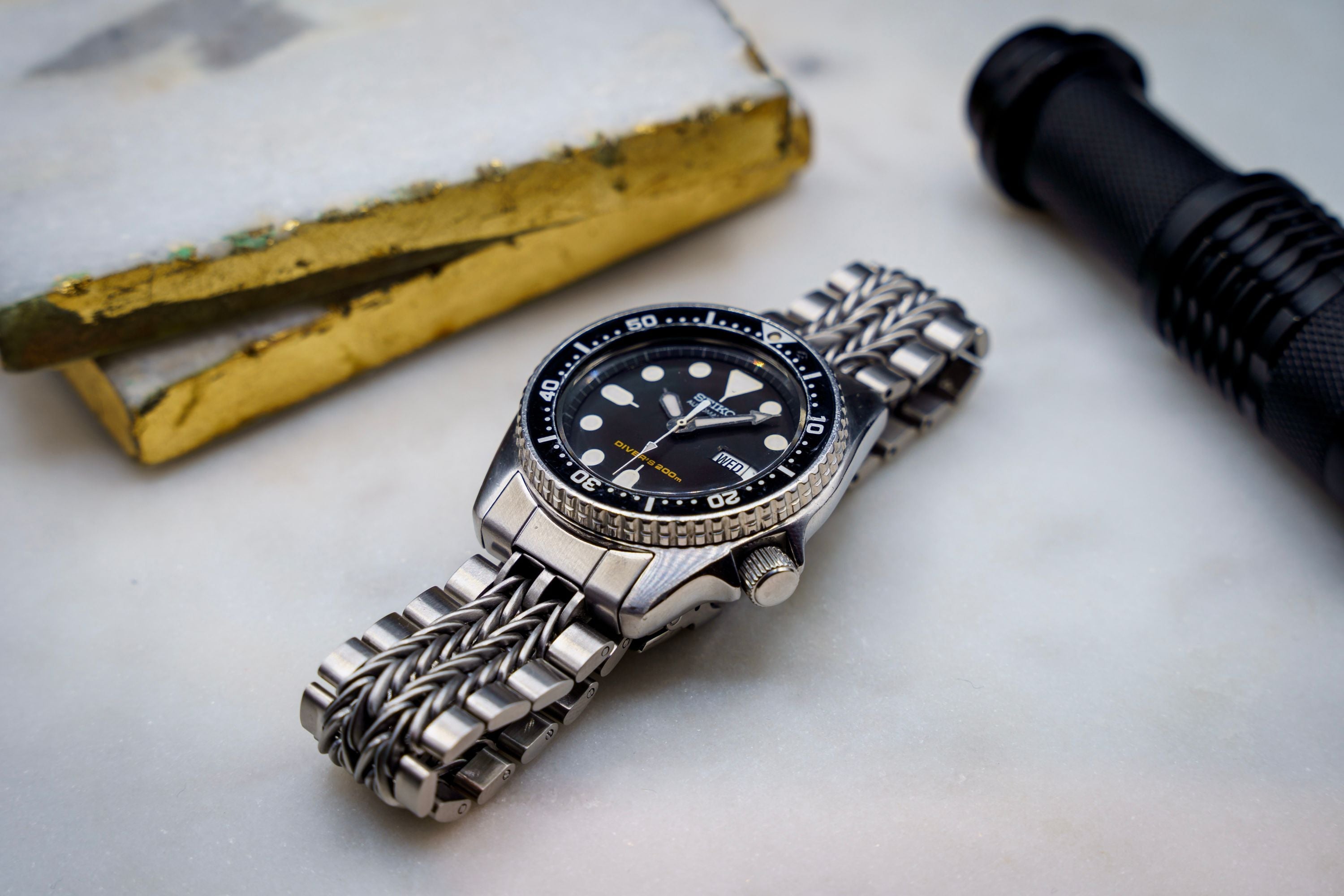 Men's diver watch with black dial and stainless steel bracelet, paired with yellow tactical pliers and black flashlight on white background
