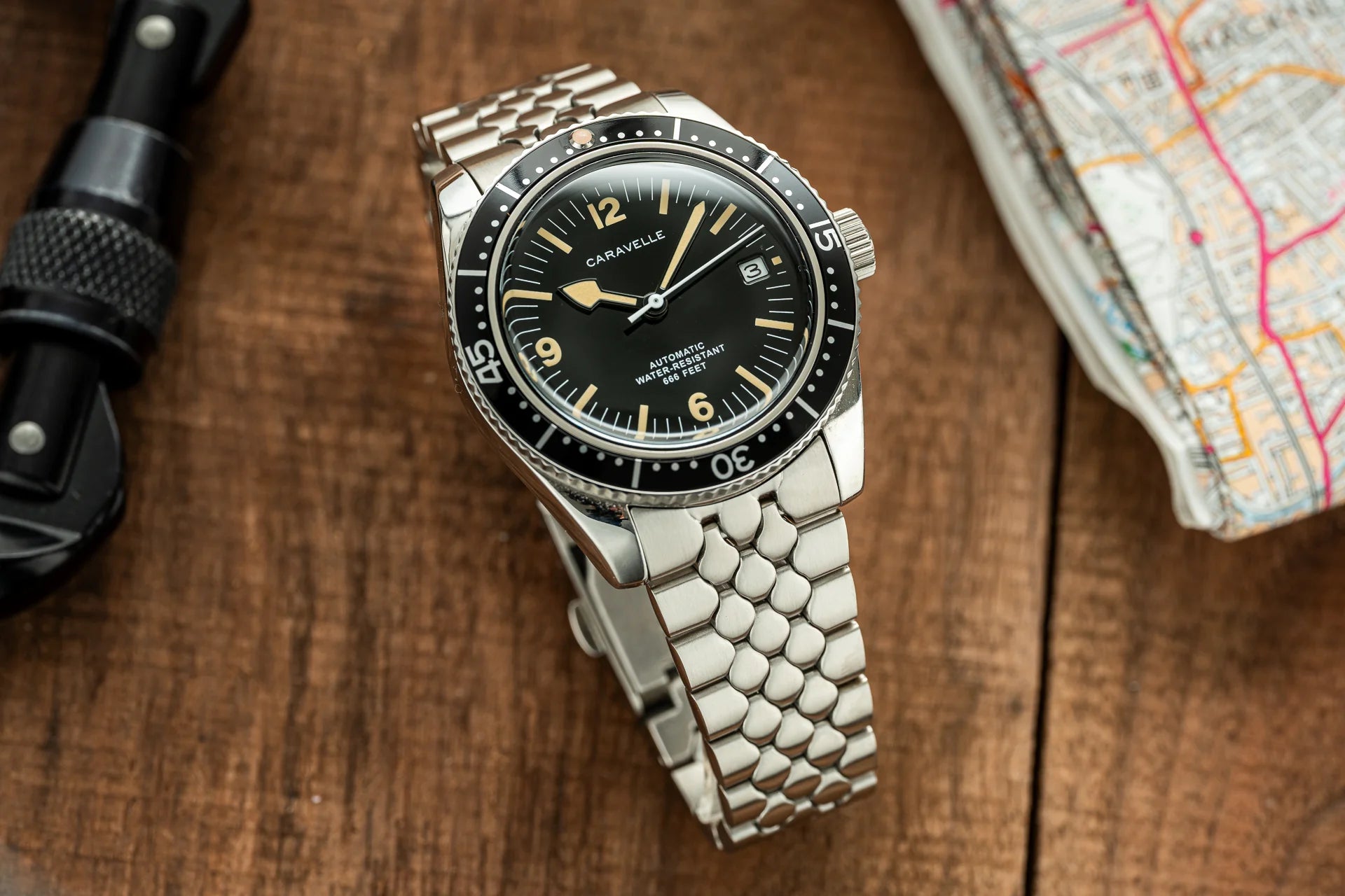 Caravelle stainless steel automatic dive watch with black dial, gold hour markers, date window and matching metal bracelet on wooden surface