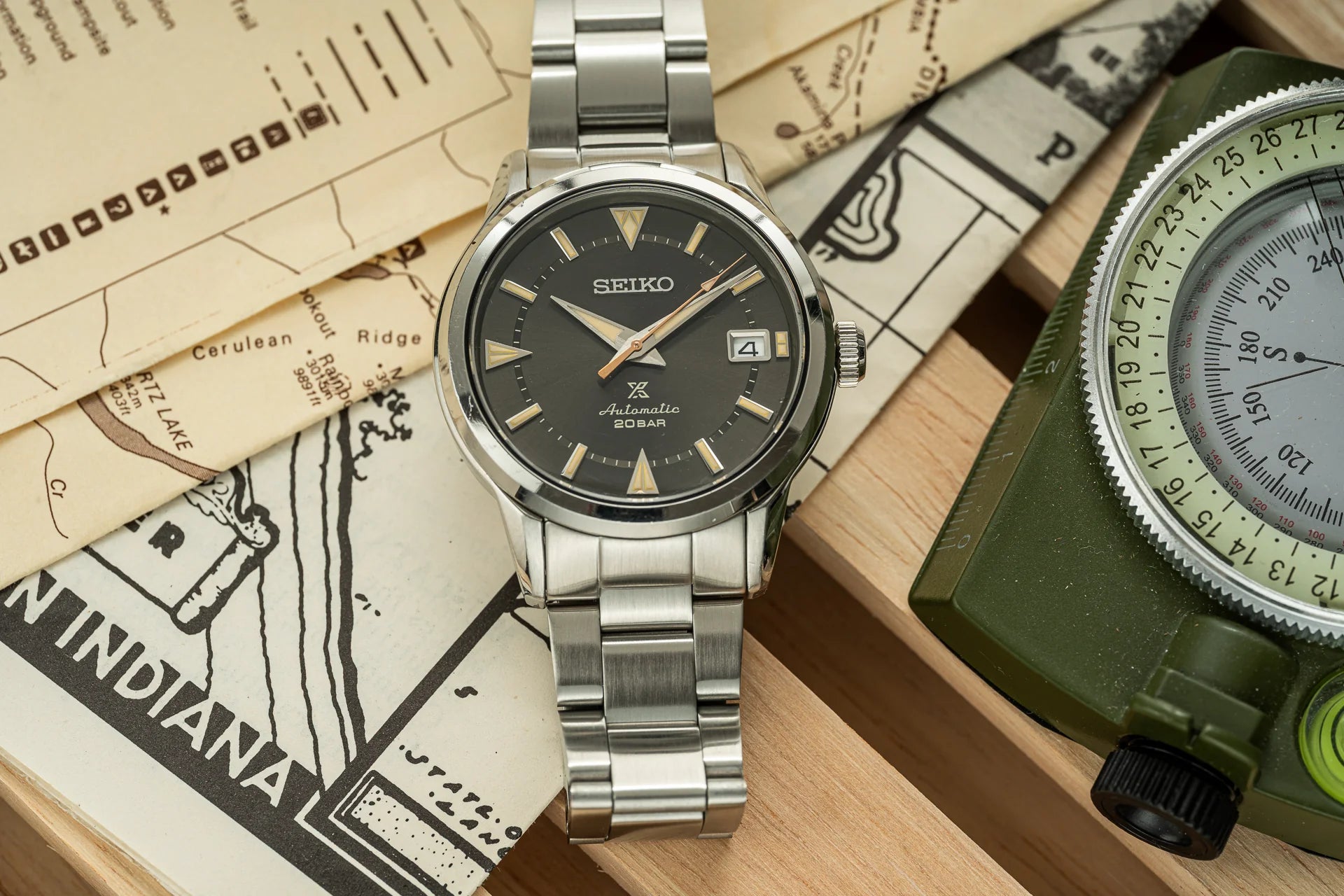 Seiko Alpinist automatic watch with black dial, stainless steel bracelet, and date window on vintage map background with compass
