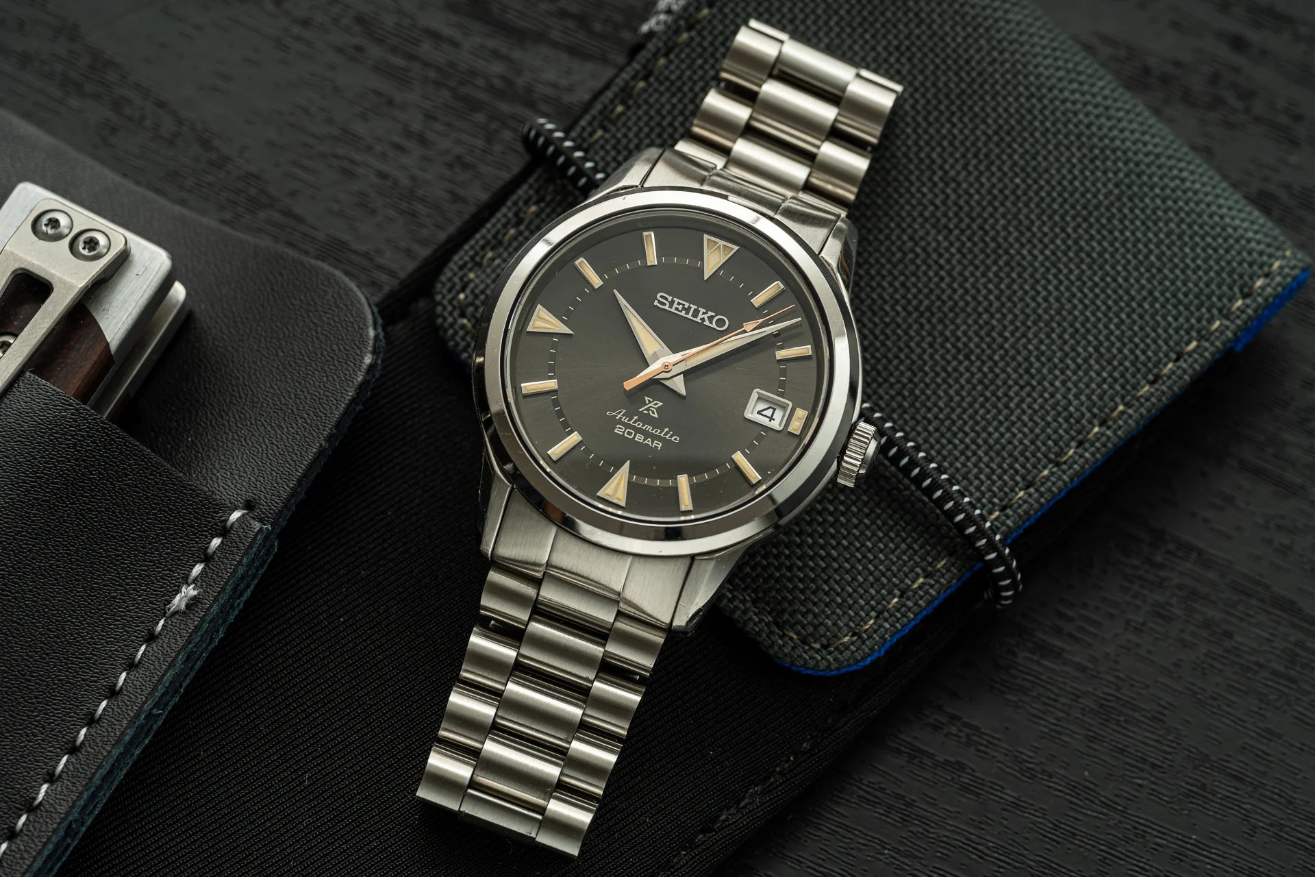 Seiko Alpinist stainless steel watch with black dial and gold indices on metal bracelet, displayed on dark fabric with accessories