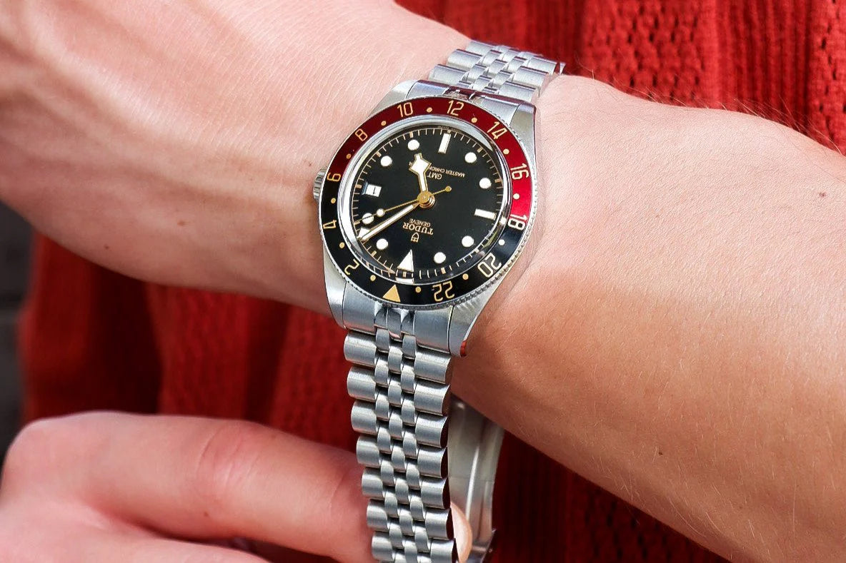 Stainless steel Tudor Black Bay 58 GMT watch with red and black bezel on jubilee bracelet, worn on wrist against red fabric background
