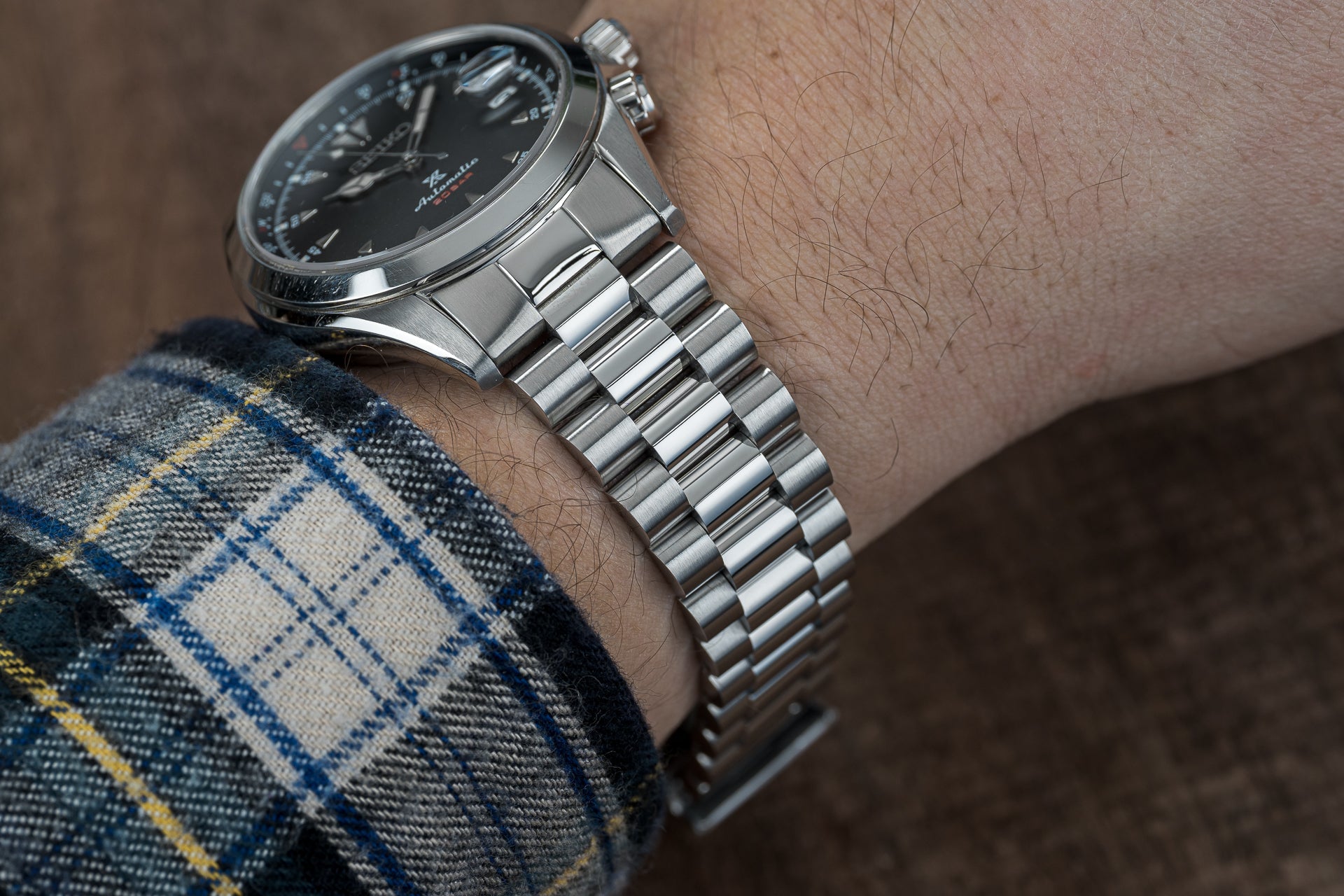 Silver stainless steel watch with black dial on three-link bracelet, worn on wrist with tartan sleeve