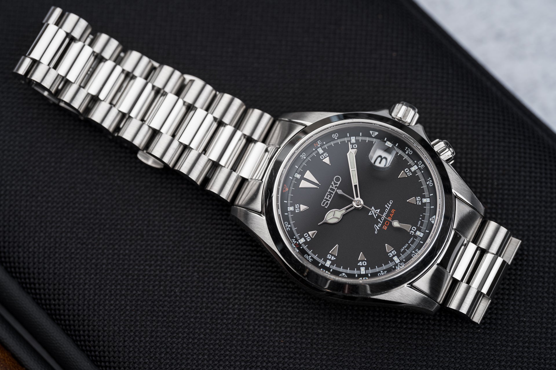 Seiko SARB017 Alpinist stainless steel watch with black dial, date window, and metal bracelet on dark fabric background