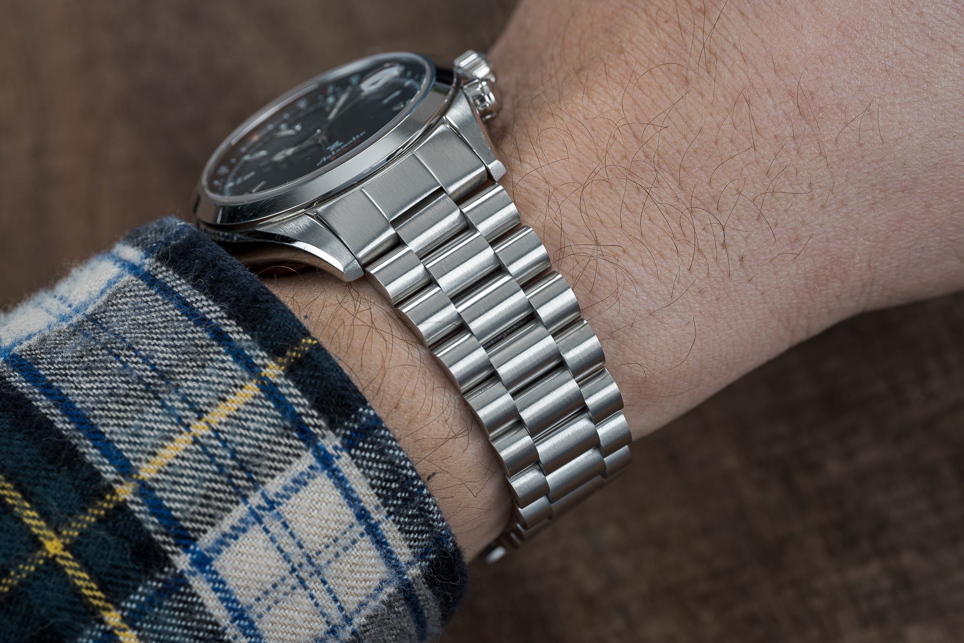 Silver stainless steel bracelet watch with black dial worn on wrist with plaid sleeve, featuring metal link band and chrome case