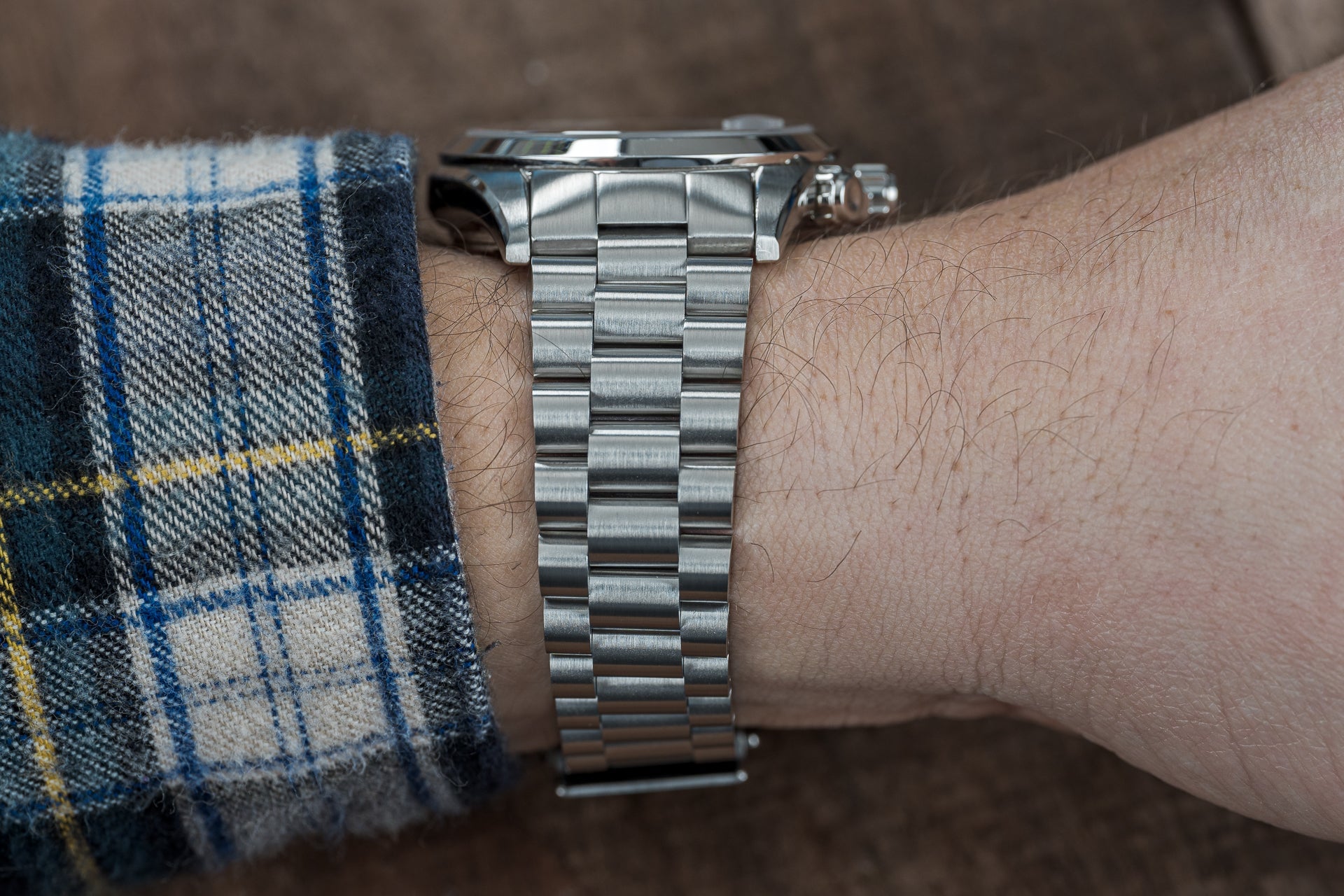 Stainless steel watch bracelet with solid links worn on wrist with plaid sleeve, showing brushed metal finish and secure clasp