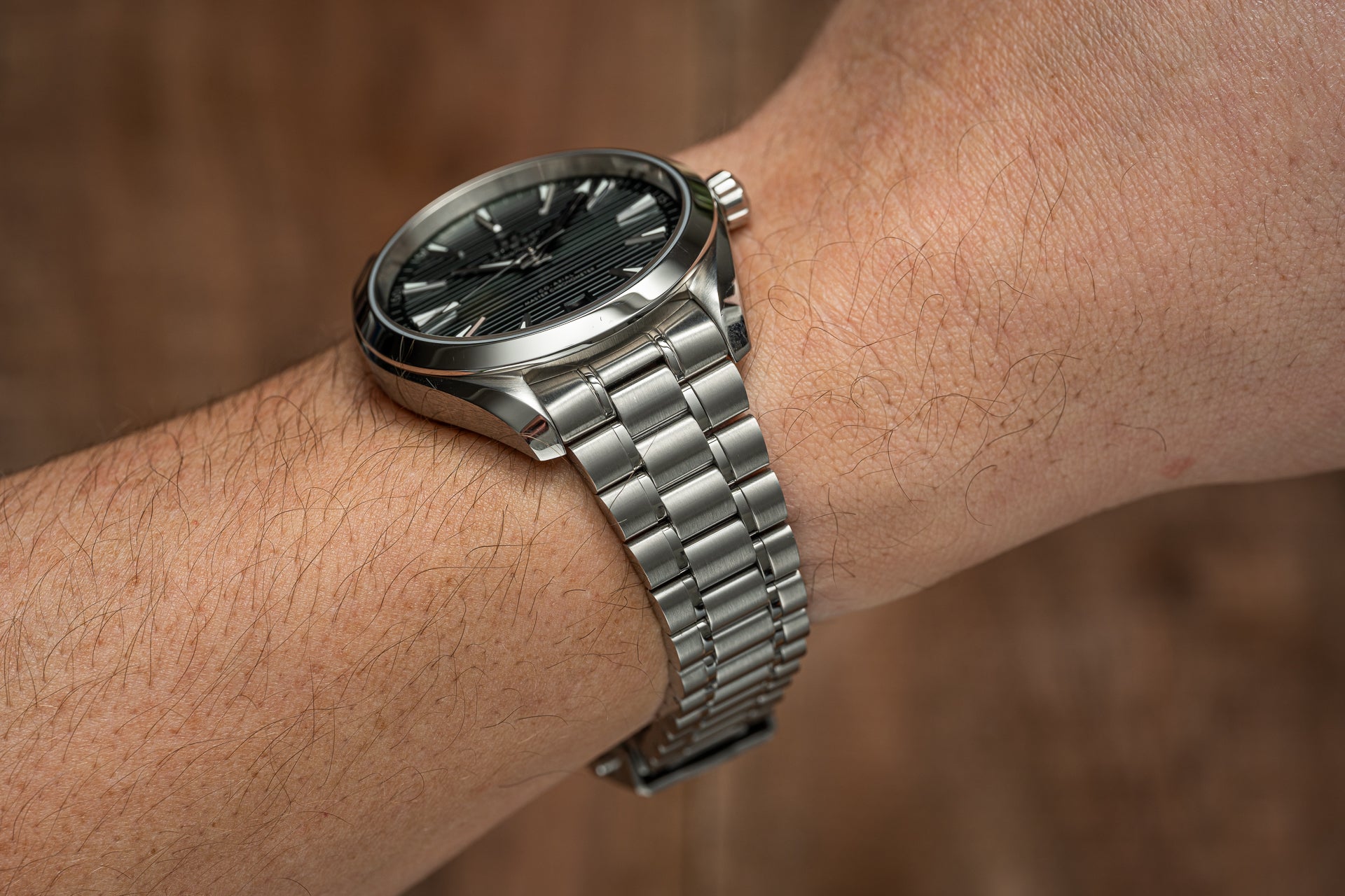Stainless steel bracelet watch with black dial and linear texture, worn on wrist against brown background