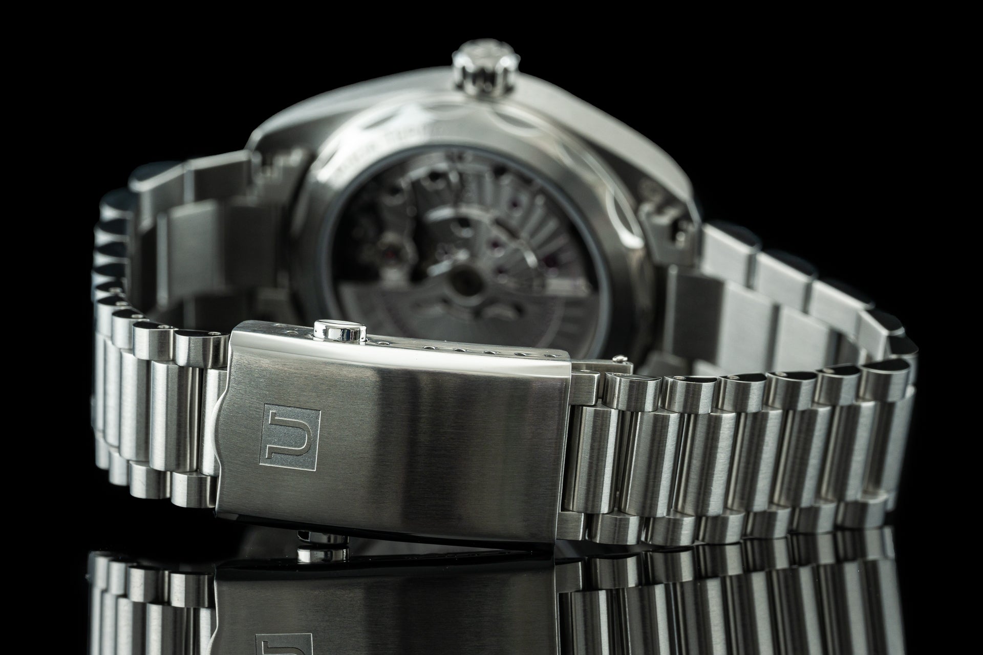 Stainless steel automatic watch with visible skeleton caseback, brushed metal bracelet with deployment clasp, and round polished case on black background