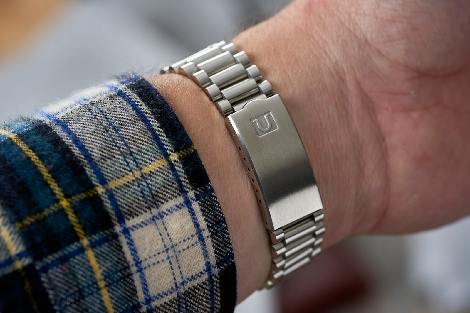 Silver stainless steel watch bracelet with cylindrical links worn on wrist, shown with plaid fabric sleeve detail
