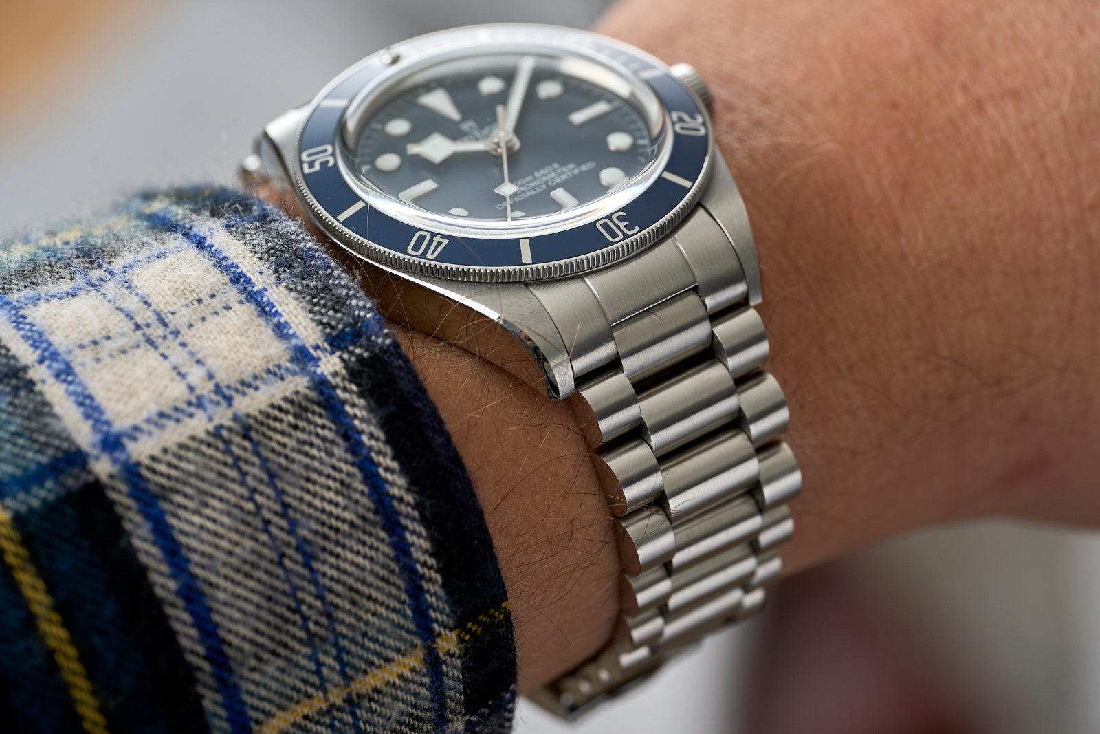 Stainless steel dive watch with black dial and blue bezel on matching metal bracelet, worn on wrist with plaid fabric sleeve