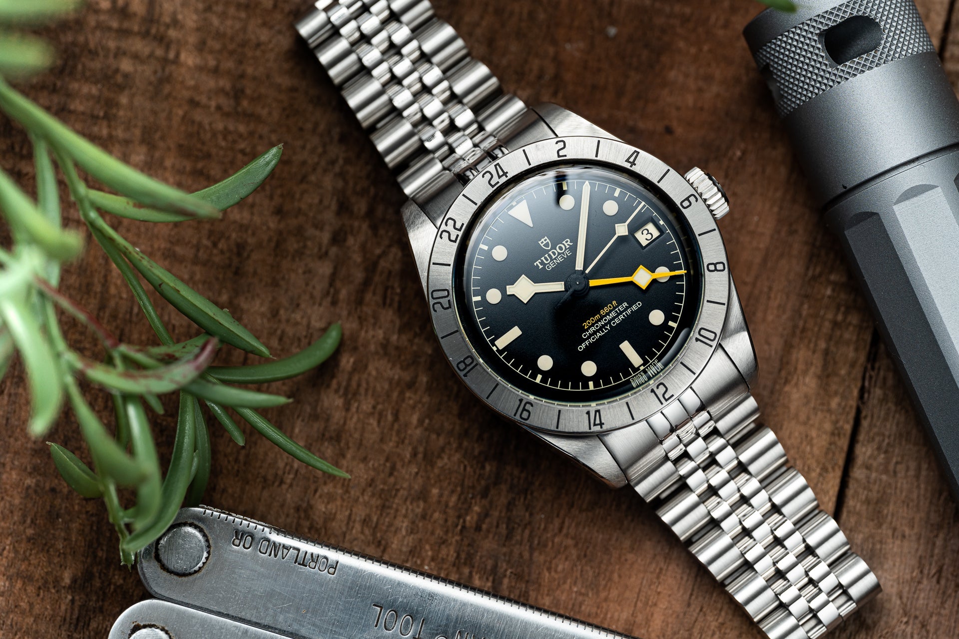 Tudor Black Bay Pro stainless steel dive watch with black dial and yellow hand on steel bracelet, displayed with outdoor gear