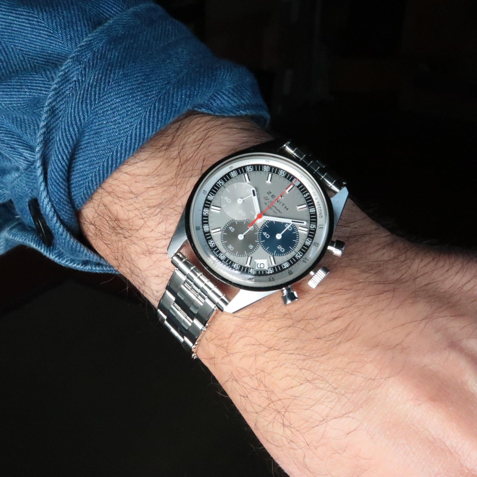 Men's stainless steel chronograph watch with silver dial and blue subdials worn on wrist with blue denim sleeve and 5-link rivet bracelet