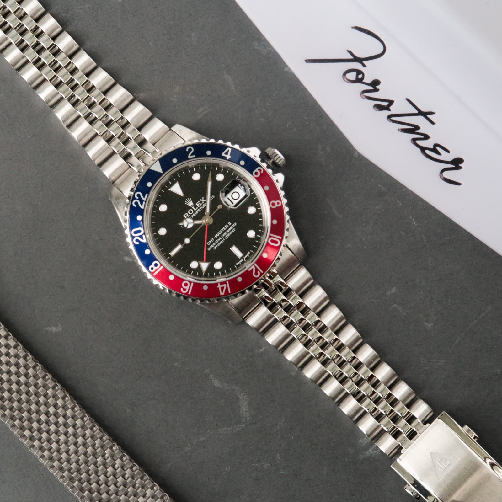Stainless steel bracelet watch with blue and red GMT bezel, black dial, and Oyster band on gray background