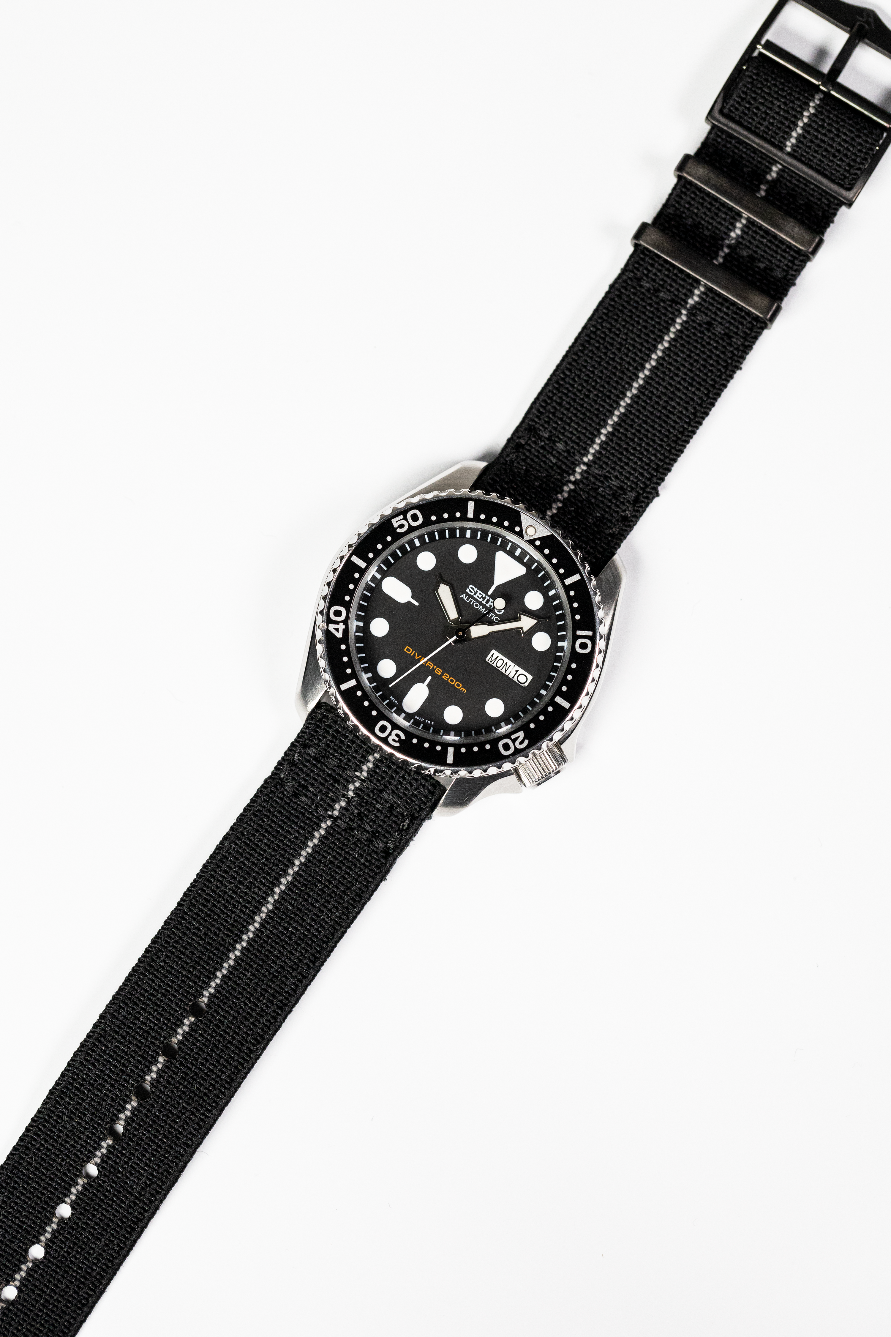 Black dive watch with stainless steel case, black dial, white dot markers, date window, and black fabric strap with white stitching