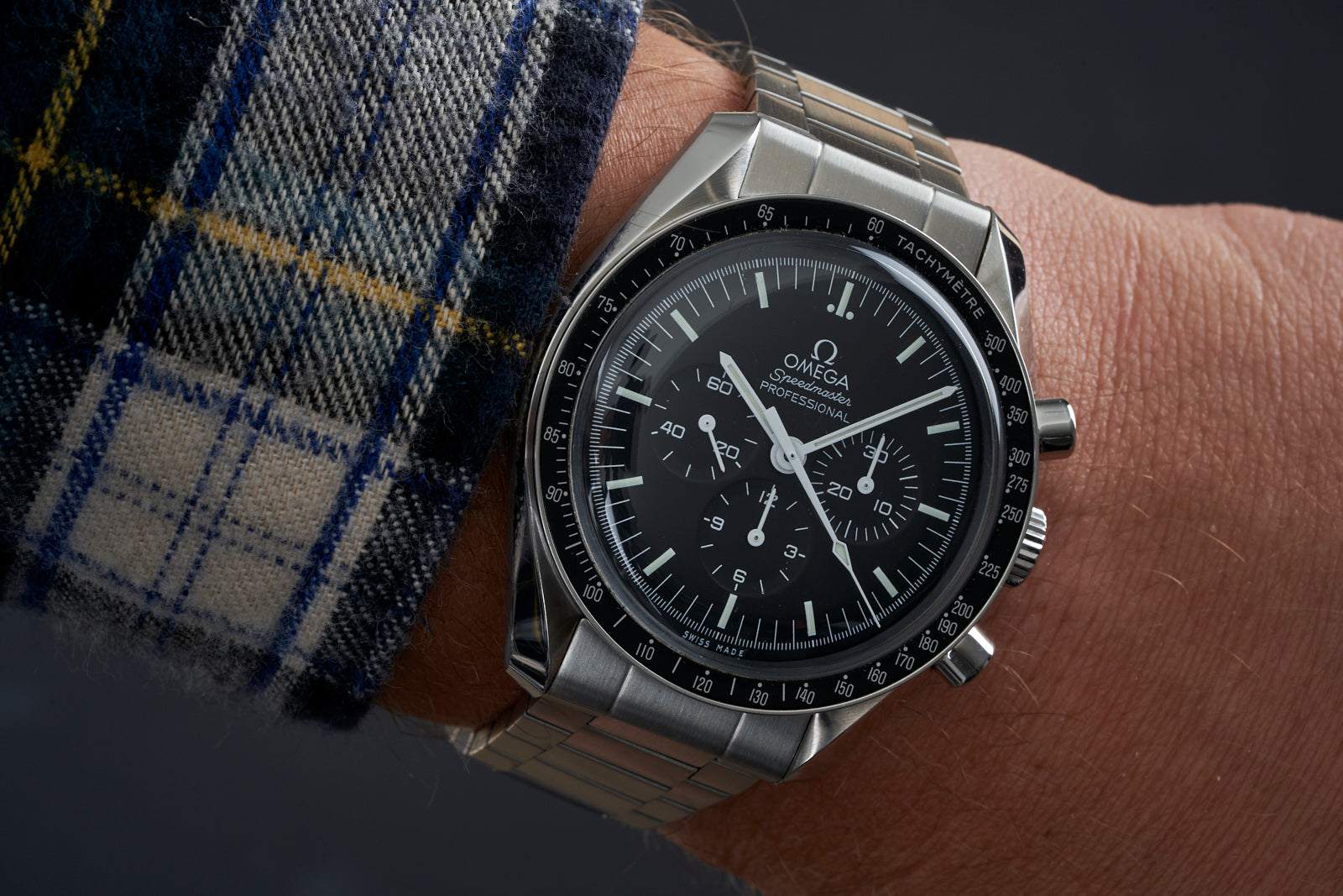 Omega Speedmaster Professional chronograph watch with stainless steel bracelet and black dial worn on wrist with plaid fabric sleeve
