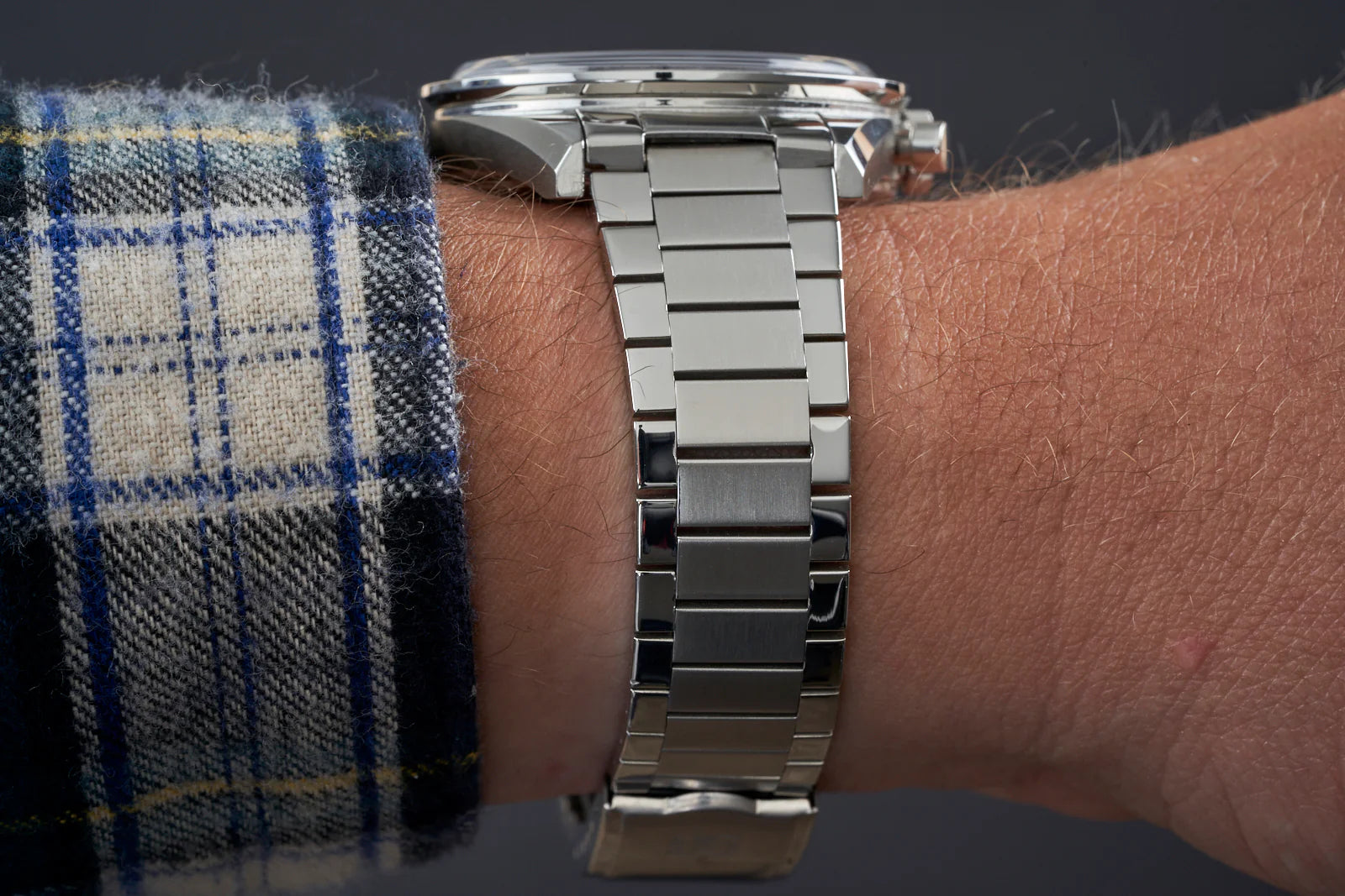 Stainless steel flat link bracelet on wrist with blue checkered shirt cuff, showing polished metal links and professional watch styling