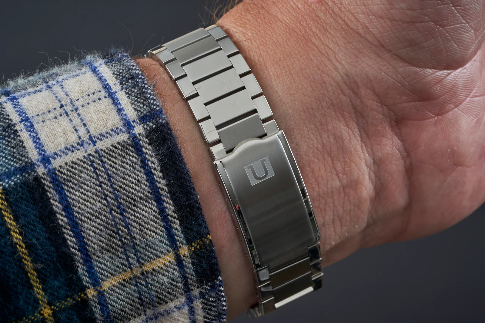 Stainless steel flat link bracelet worn on wrist with plaid shirt cuff, showing polished metal finish and secure clasp closure