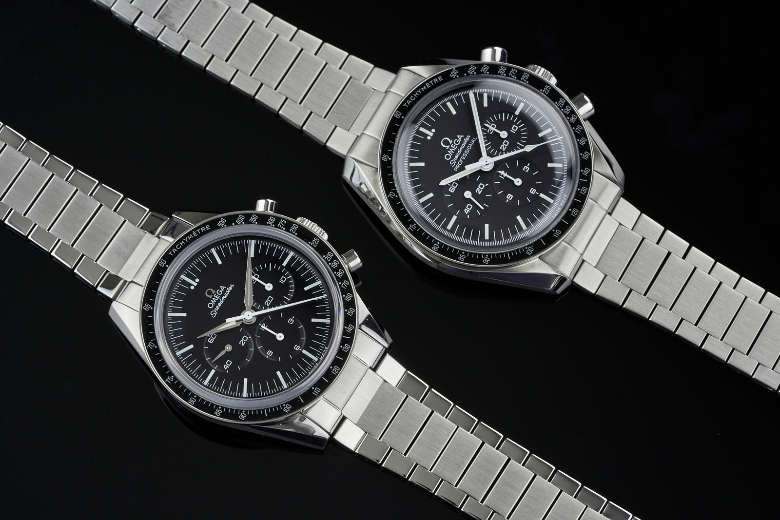 Pair of stainless steel chronograph watches with black dials, tachymetre bezels, and flat link metal bracelets on black background