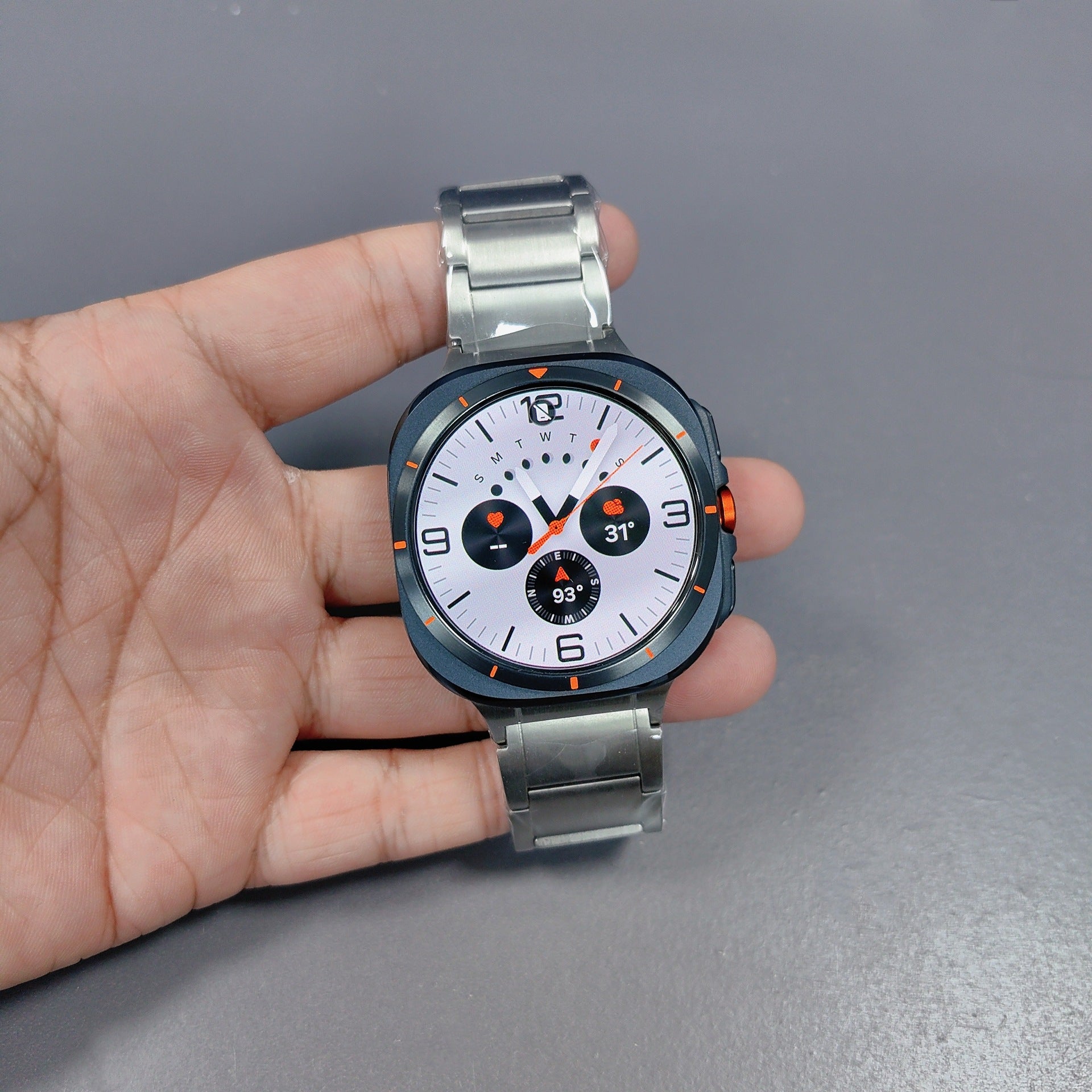 Smartwatch with titanium bracelet held in hand, white dial face with black chronograph subdials, orange accents and digital display showing heart rate and temperature