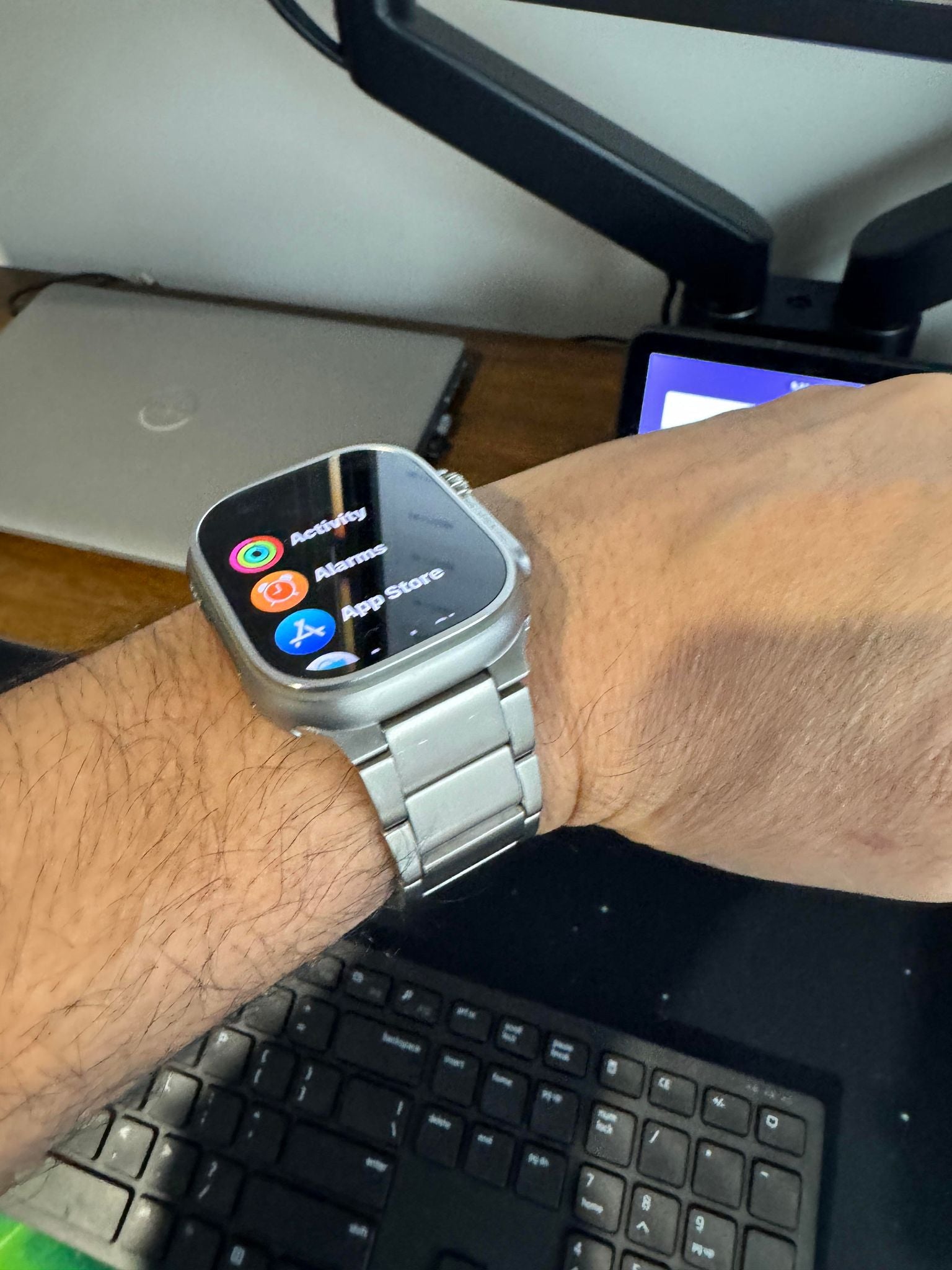 Silver titanium smartwatch with square display showing app menu, worn on wrist with metal band bracelet over keyboard desk setup