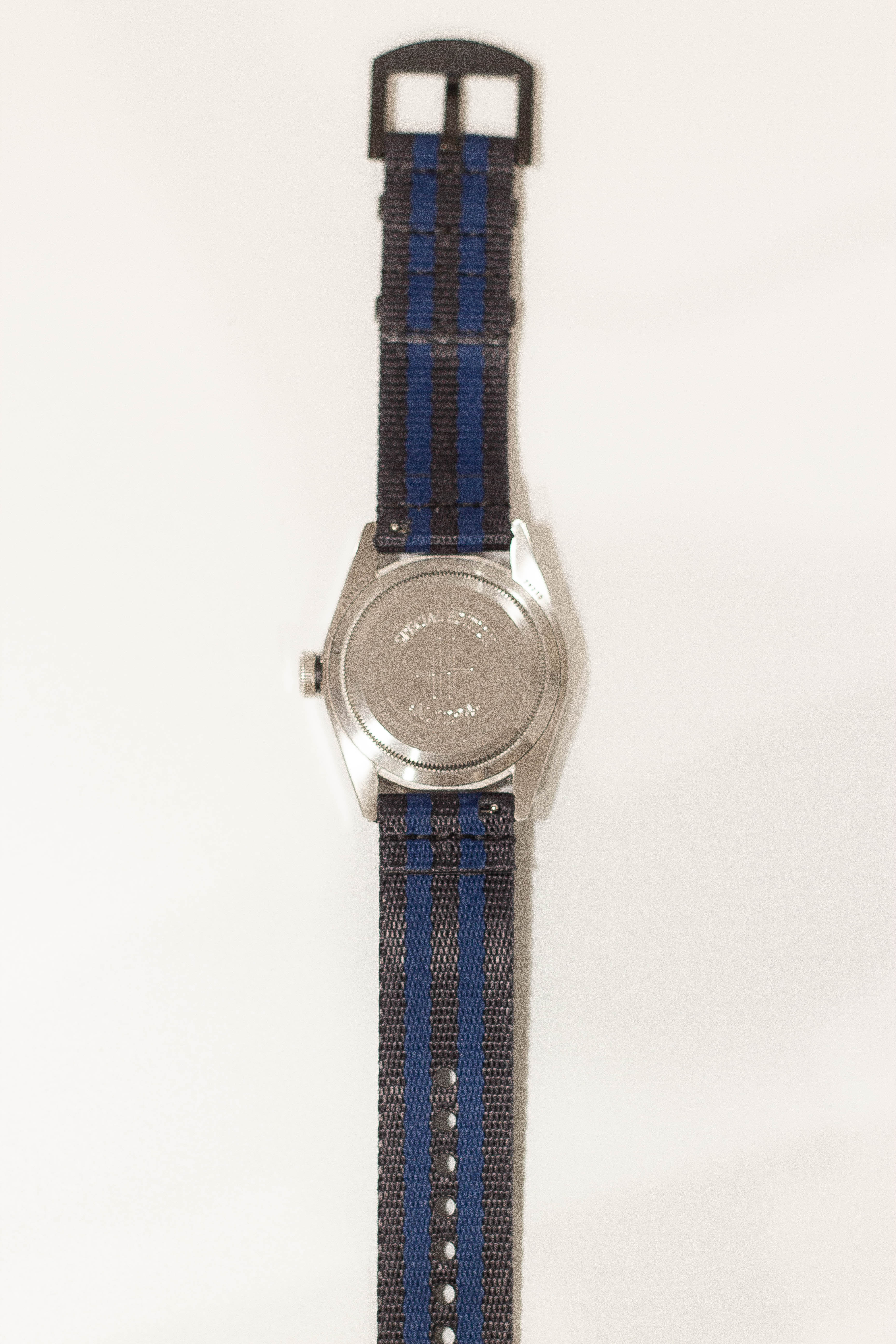 Stainless steel watch with blue and black striped fabric band and black buckle hardware on beige background
