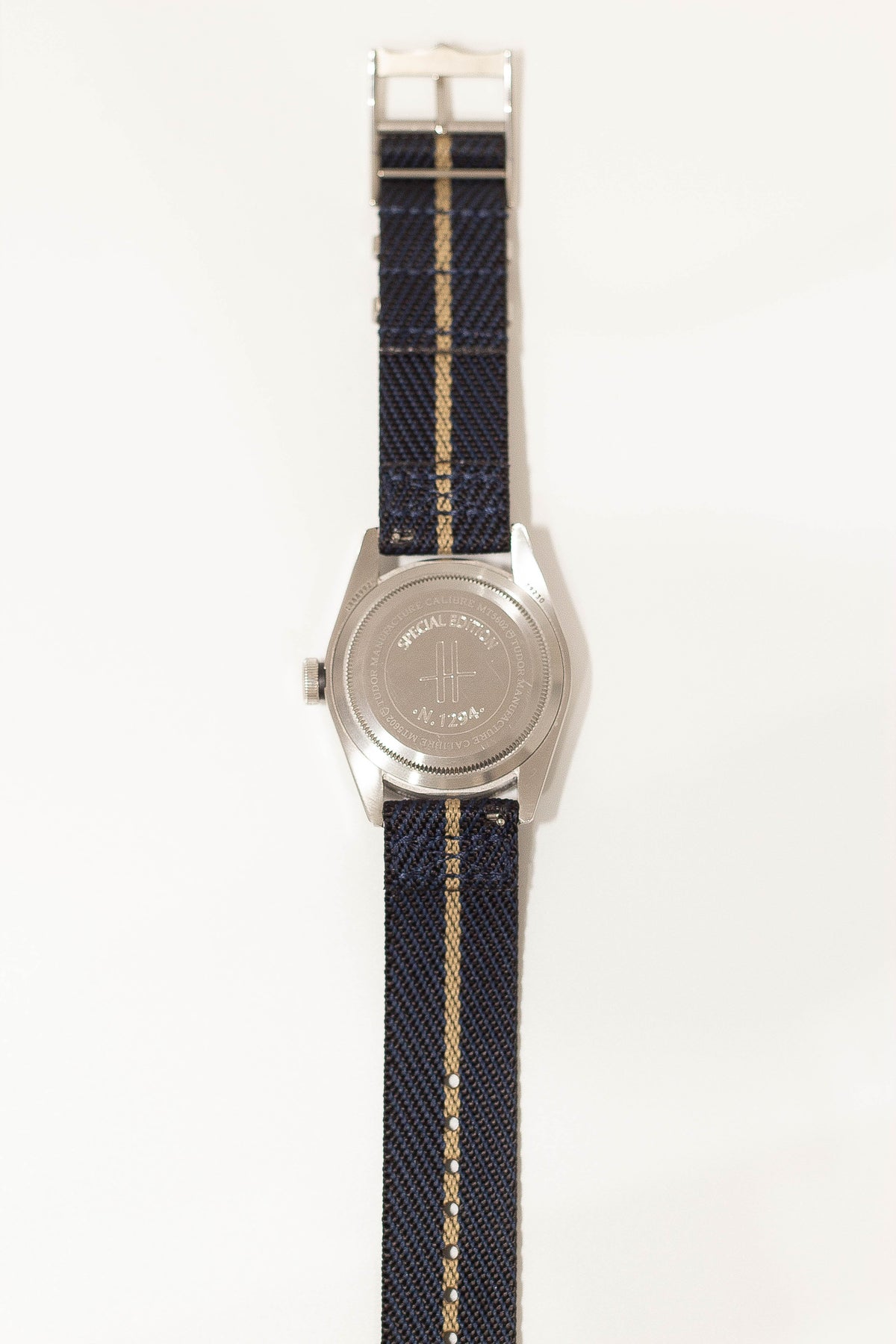 Military-style watch with stainless steel case, navy canvas band with tan stripe, and engraved case back detail