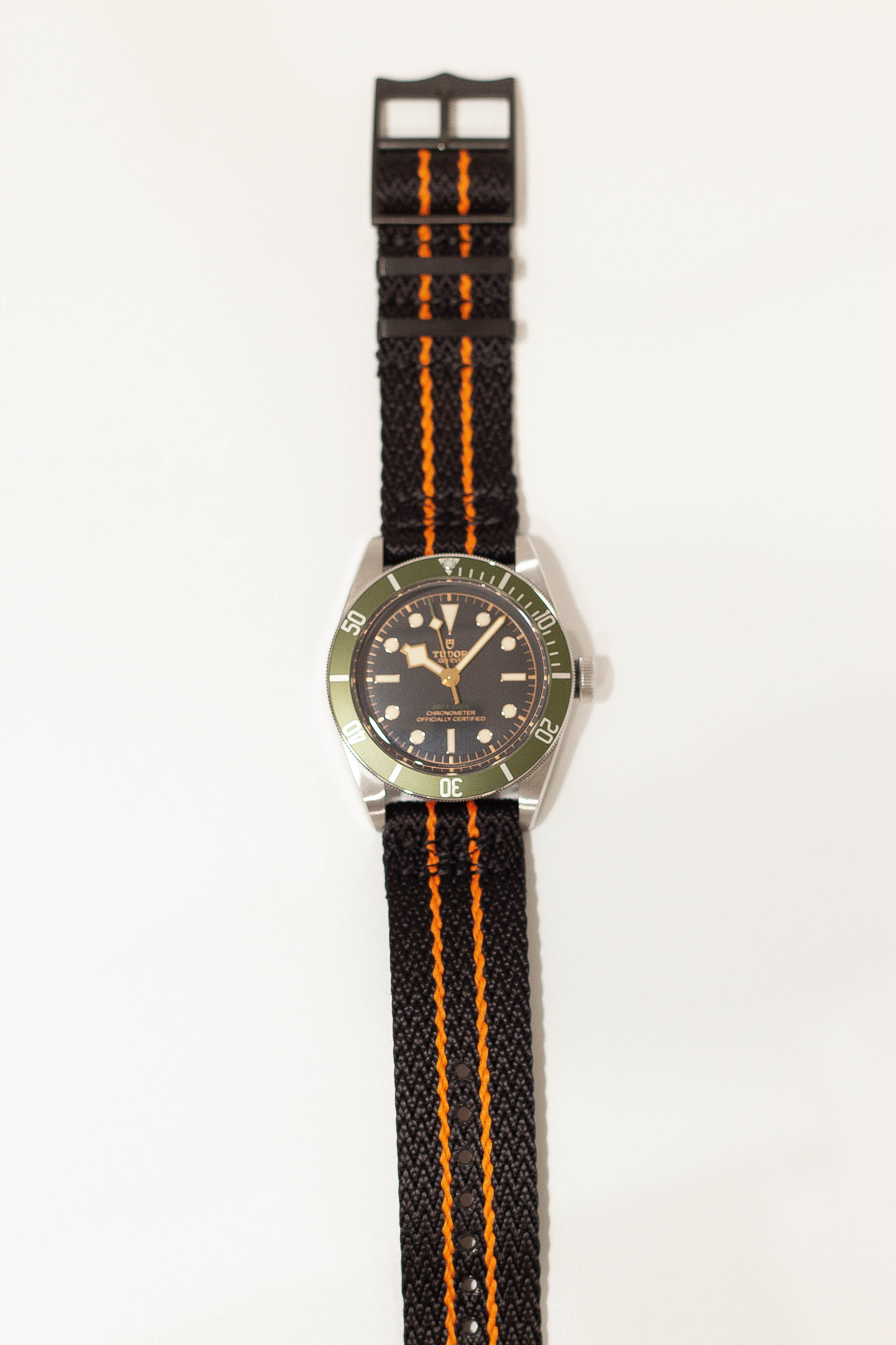 Stainless steel dive watch with olive green bezel, black dial, and black fabric strap with orange racing stripes, featuring quick release buckle