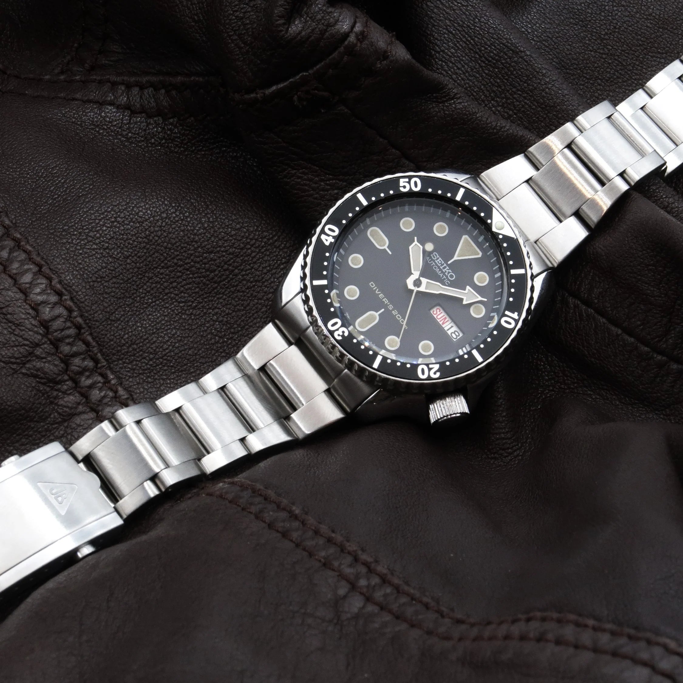 Stainless steel dive watch with black dial and bezel on metal bracelet, displayed on black leather jacket sleeve