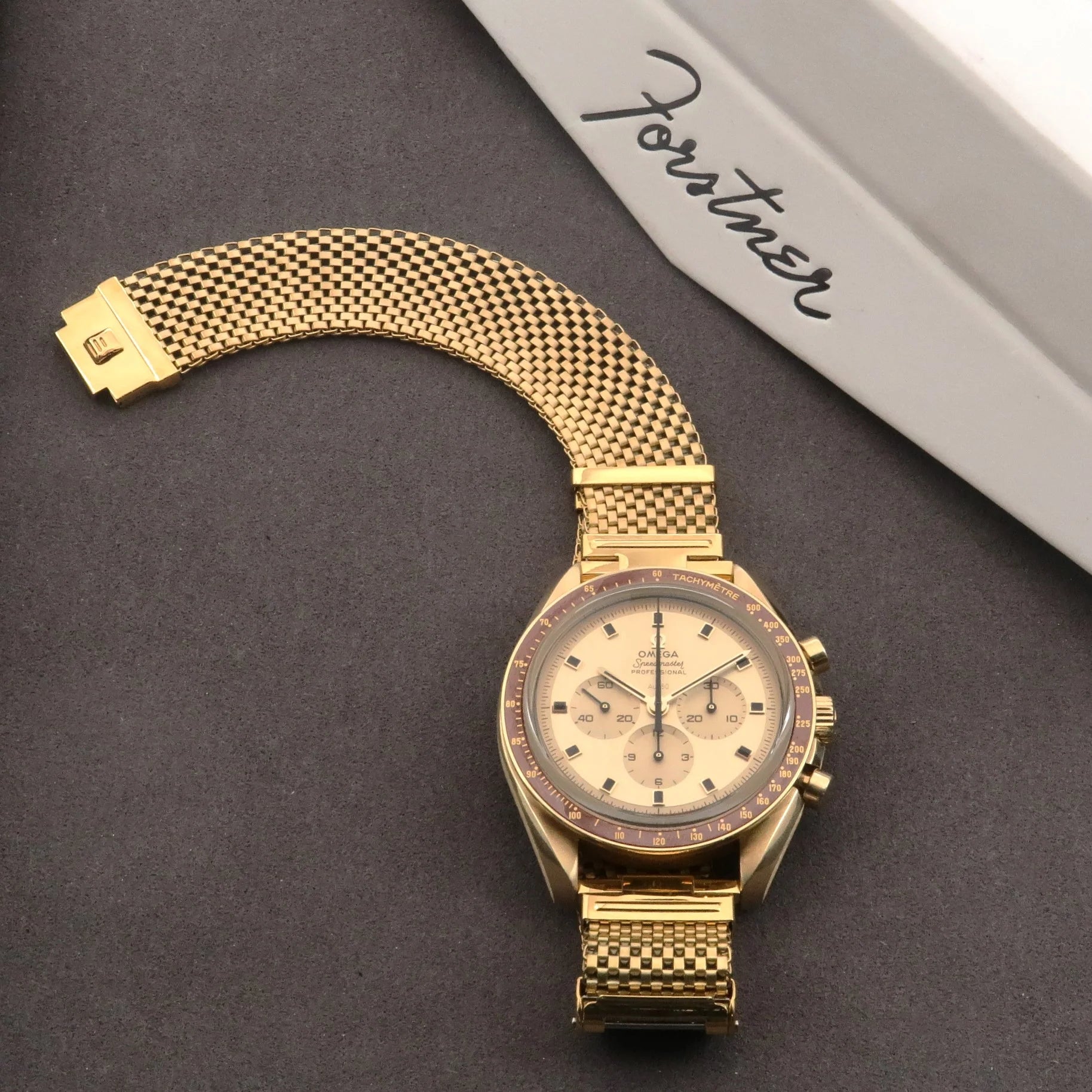 Gold plated mesh watch bracelet with straight ends and folding clasp on dark surface next to branded box