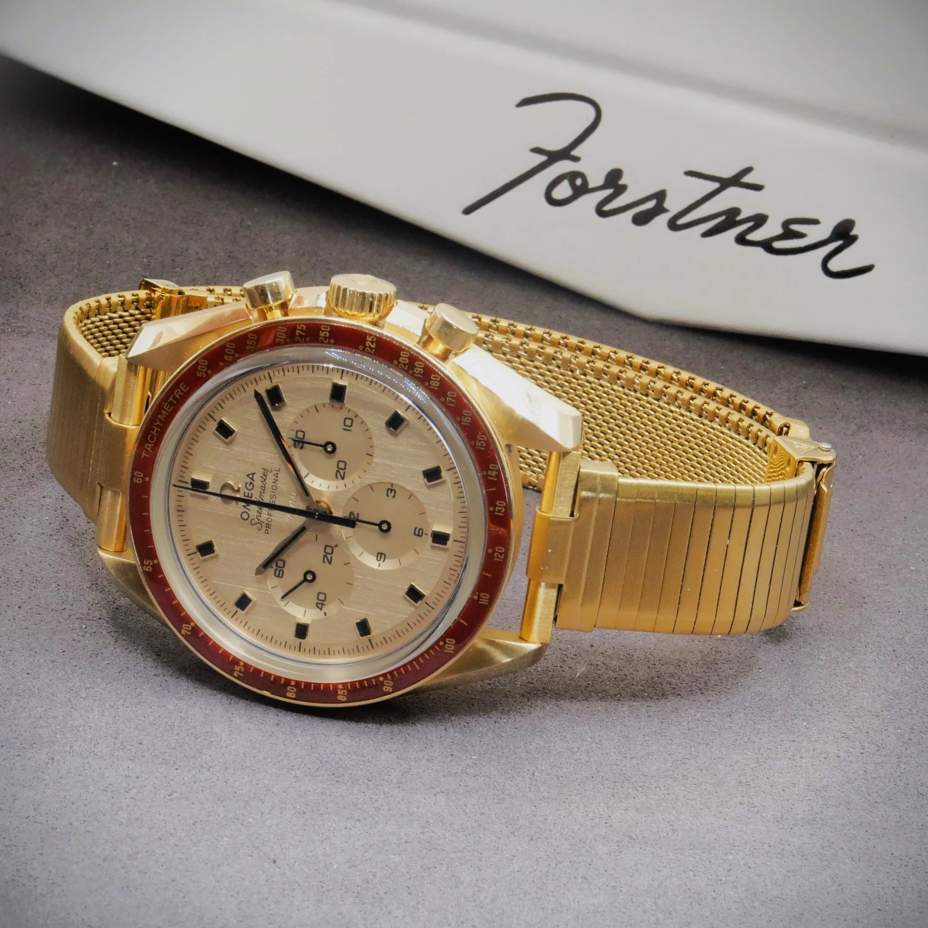 Gold plated chronograph watch with red bezel, mesh bracelet, cream dial and black subdials on gray surface