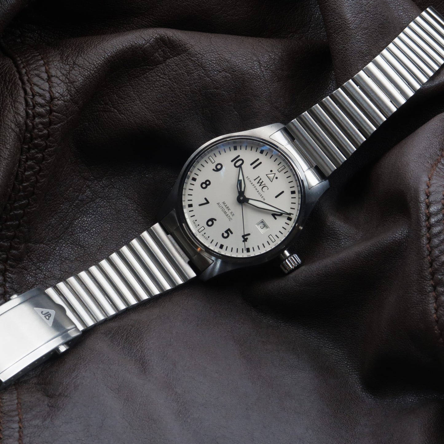 IWC Mark XX watch with stainless steel barrel bracelet on black leather background, showing white dial and date window