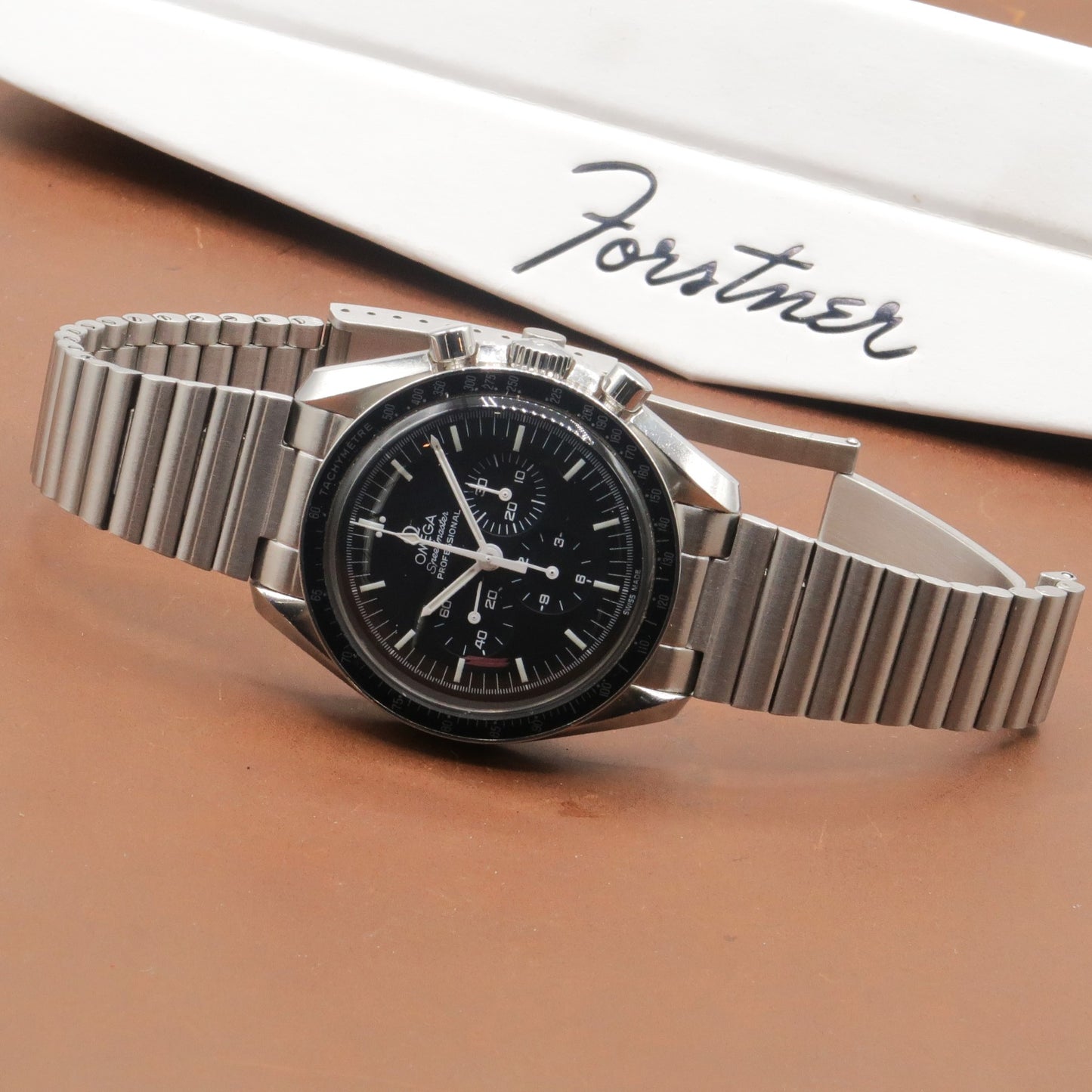 Omega Speedmaster chronograph watch with black dial and stainless steel barrel bracelet on tan background