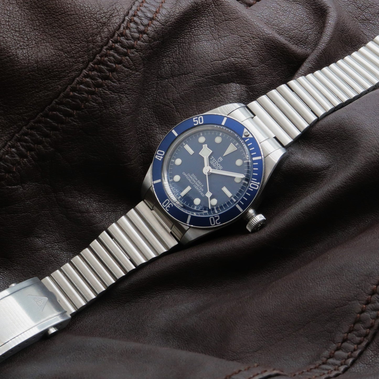 Forstner barrel bracelet steel link band on Tudor Black Bay watch with blue dial and bezel against dark leather jacket background