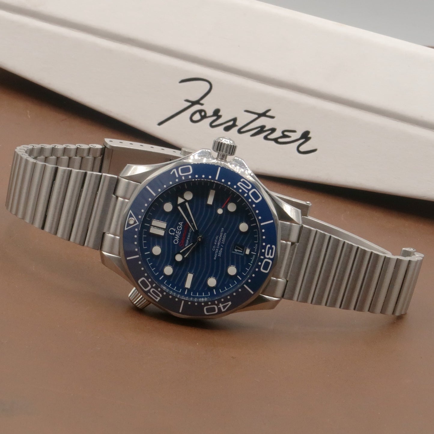 Omega Seamaster diver watch with blue dial and stainless steel barrel bracelet on tan background next to Forstner box