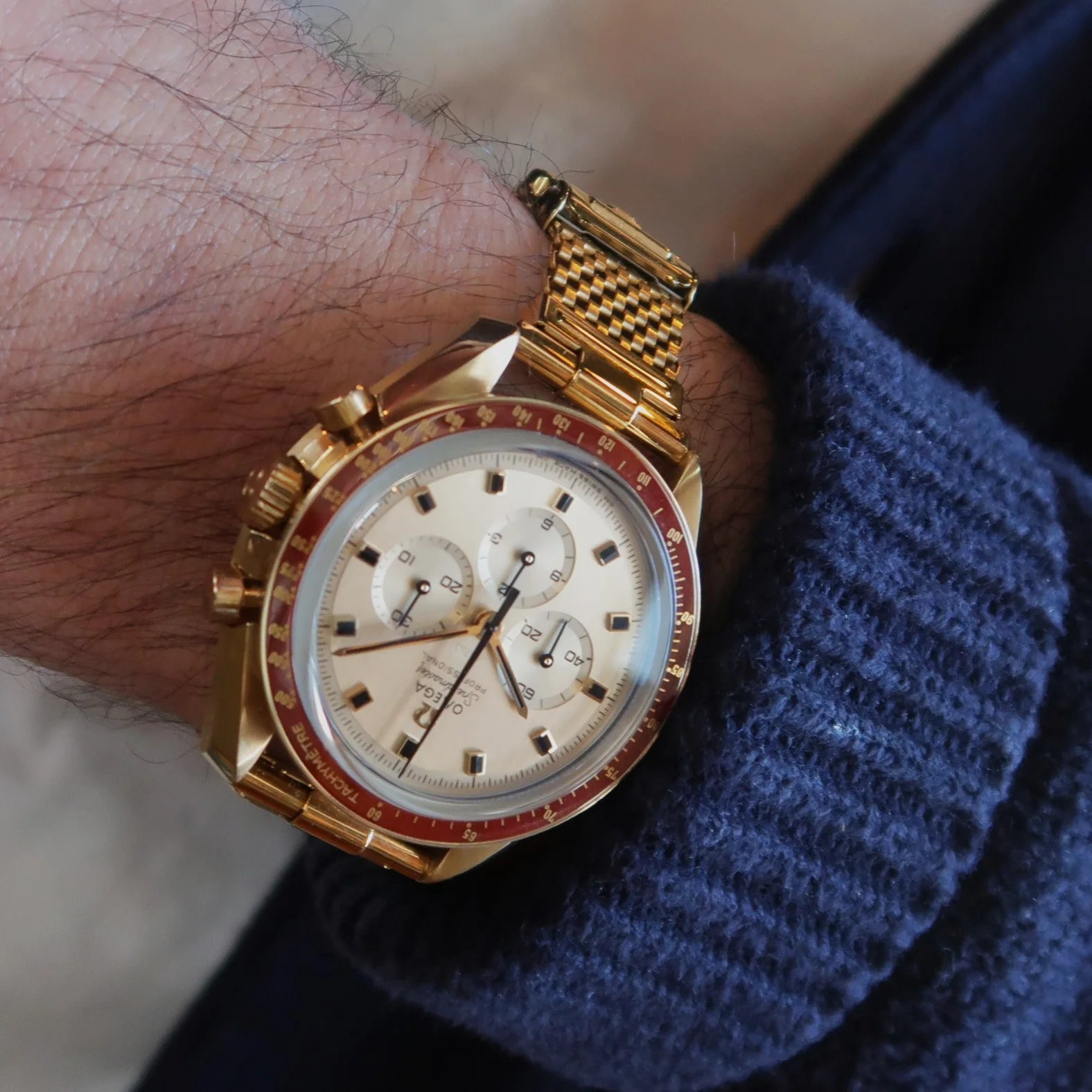 Gold plated chronograph watch with red bezel and mesh bracelet worn on wrist with blue sleeve